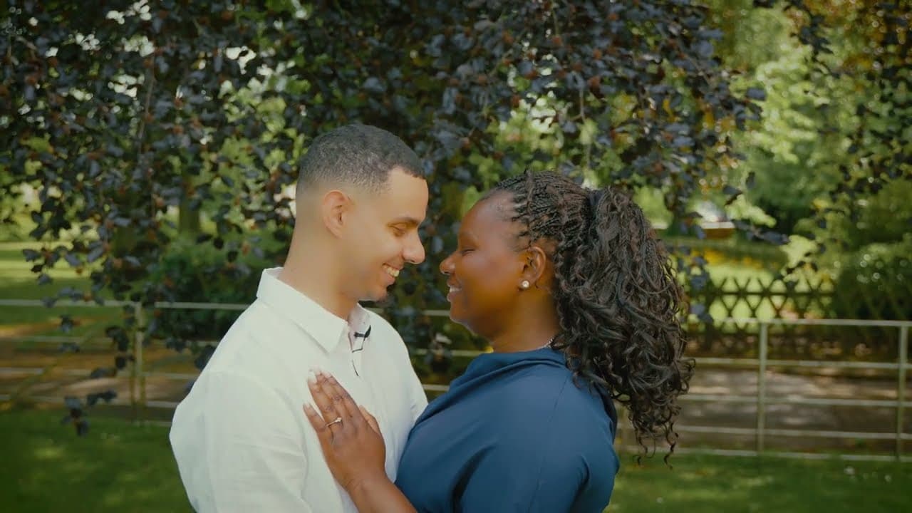 The couple face to face in a sunlit park, foreheads almost touching, her hand flat against his white shirt under a copper beech.