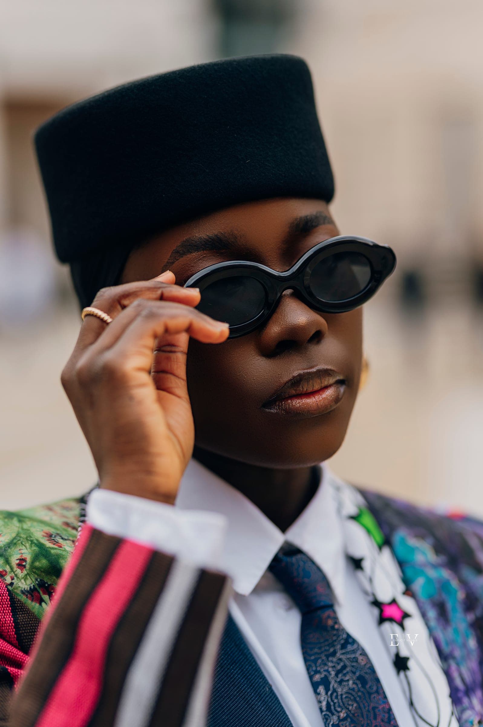 The same look with one hand raised to the frame of her sunglasses, a gold ring on her finger, the printed lapels bright at the edge of the frame.