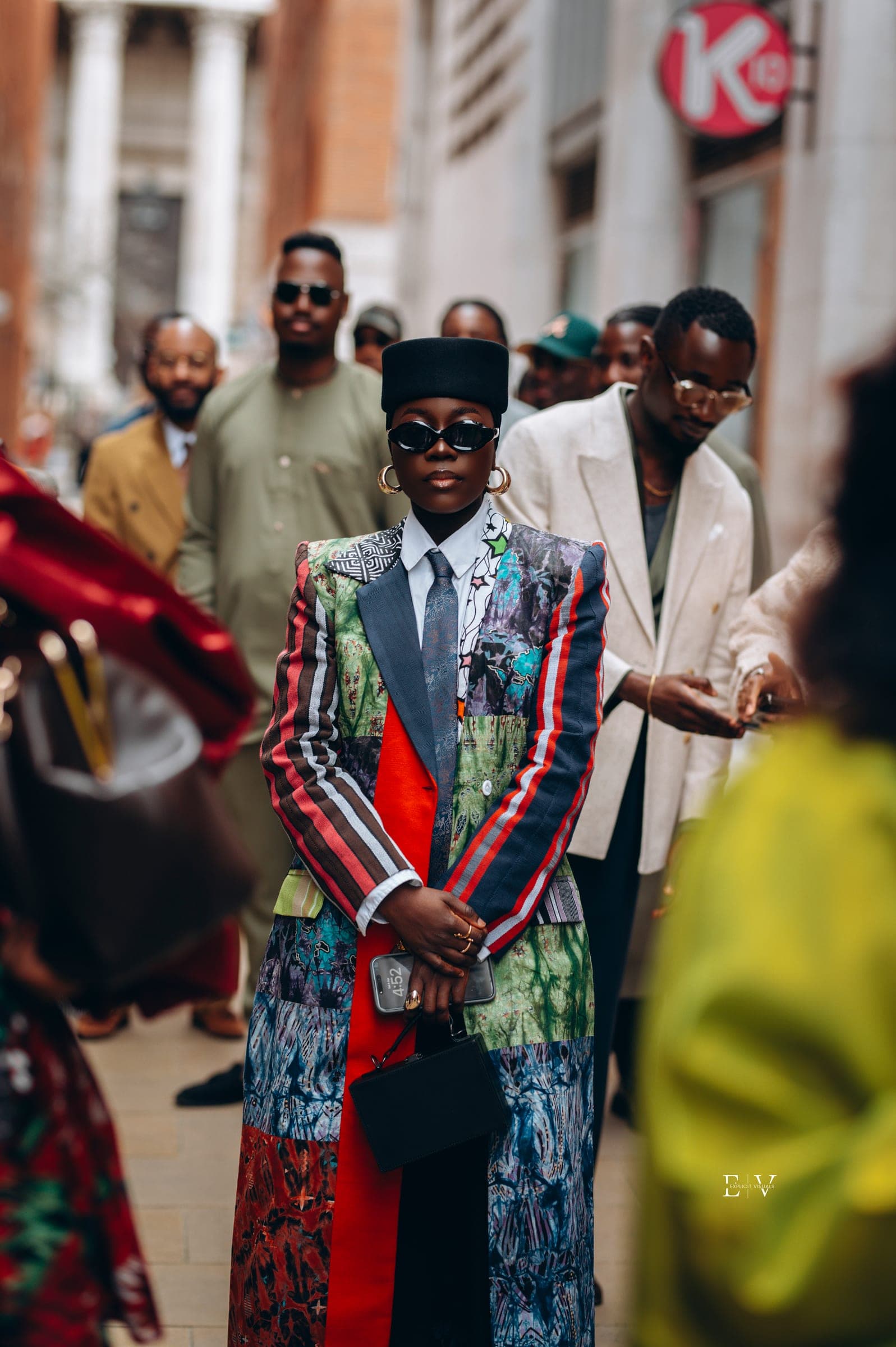 The same coat photographed from further back, the wearer walking through a busy street with other guests blurred either side.