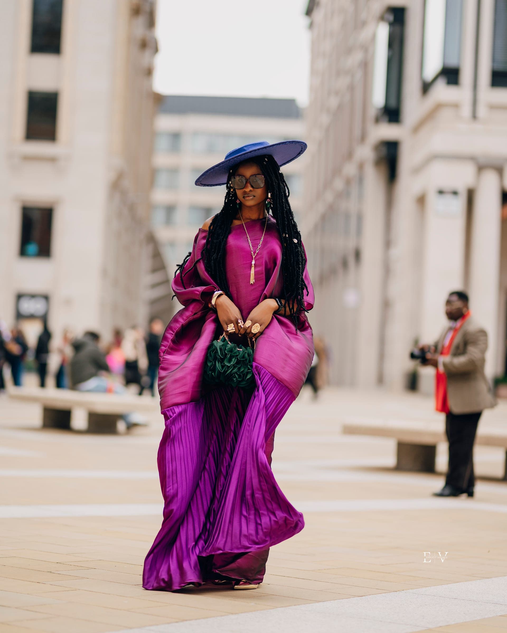 The woman in the magenta gown mid-stride, the pleated skirt swinging out, a green woven bag held in both hands and a wide blue brim shading her face.