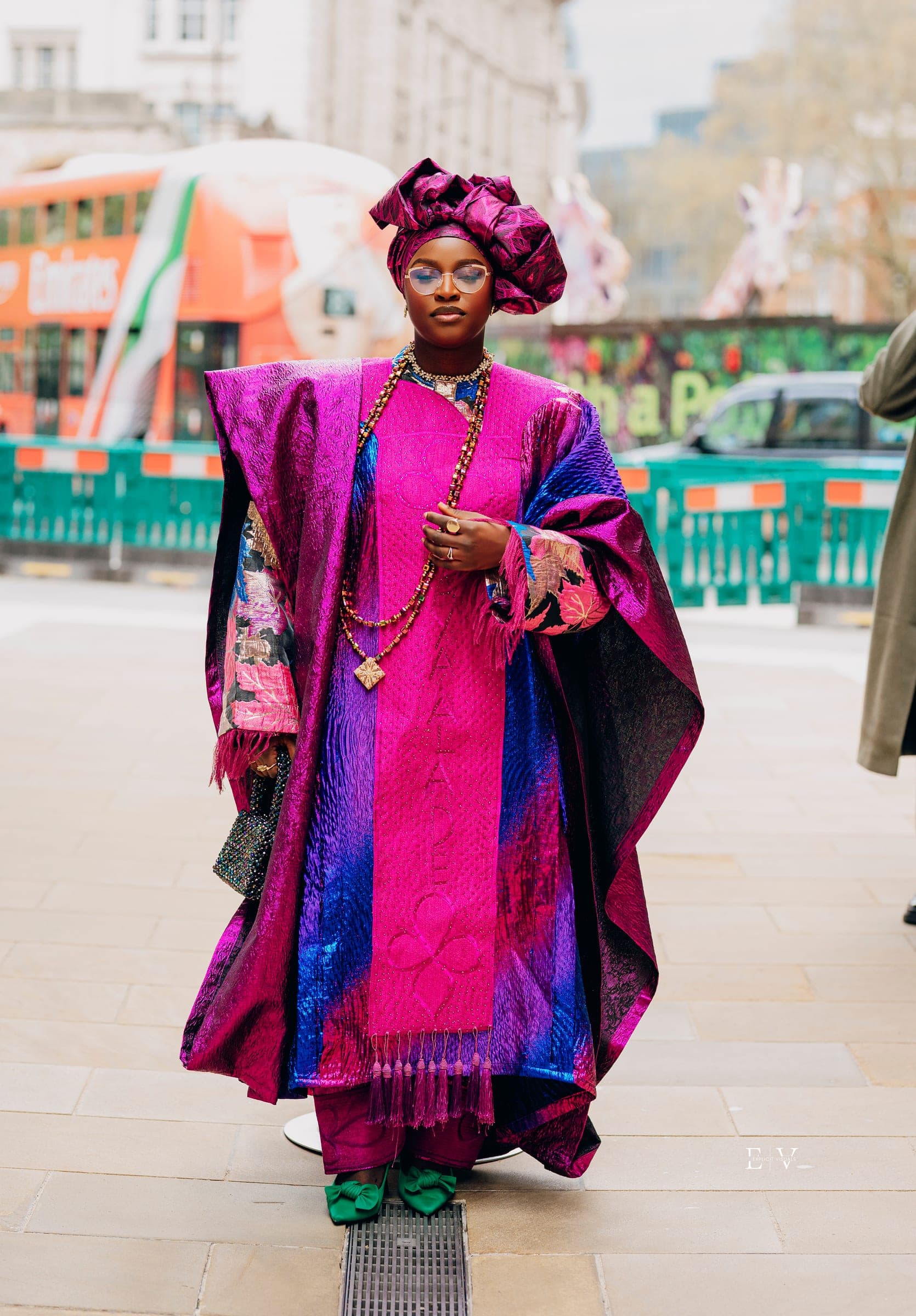A woman in a fuchsia and purple agbada with a matching gele and layered amber beads, standing on a London street with a red bus and traffic barriers behind.
