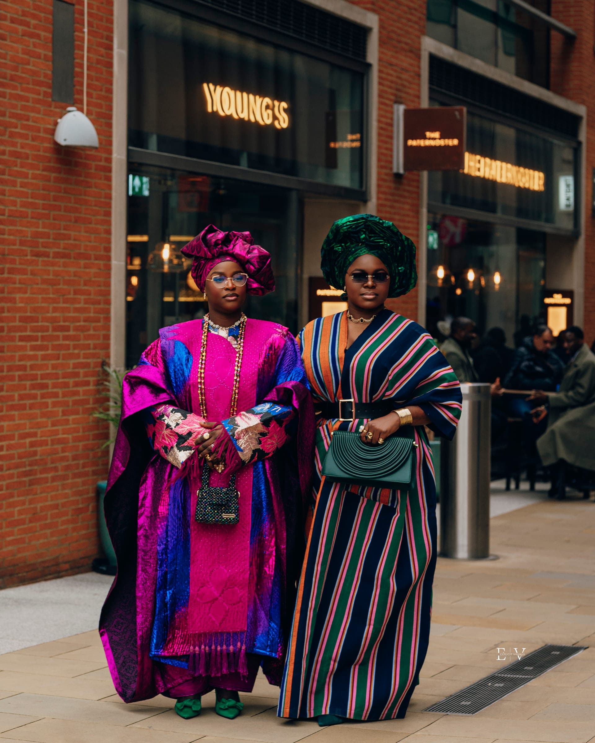 Two women side by side outside a brick-fronted pub — one in a magenta and blue agbada with a matching gele, one in a green gele and a striped wrap gown holding a green handbag.