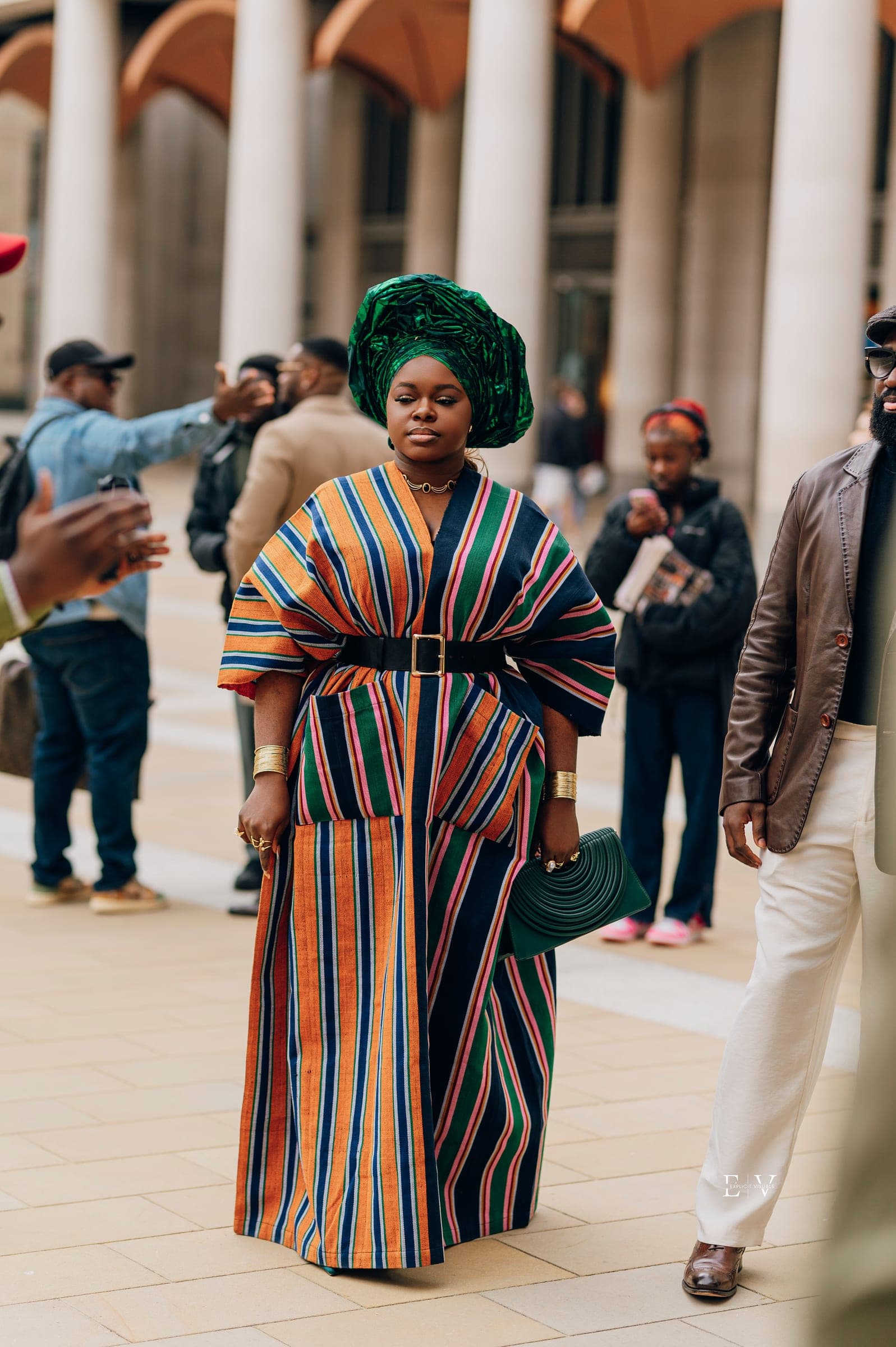 The same striped gown and green gele photographed among other guests, a gold cuff on each wrist and a green bag at her side.