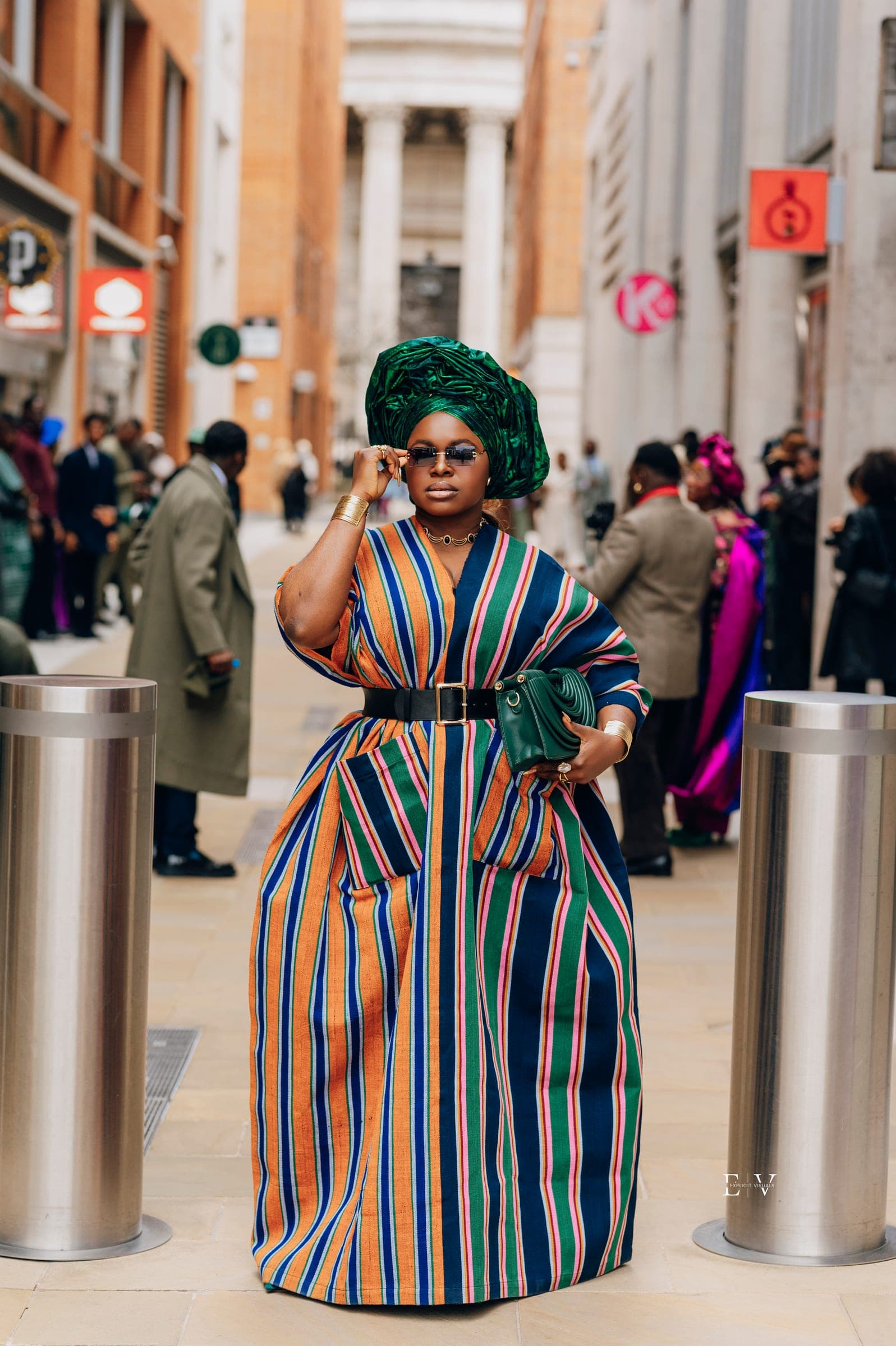 Standing between two steel bollards on the street, one hand lifted to her sunglasses, the striped skirt falling full to the ground.
