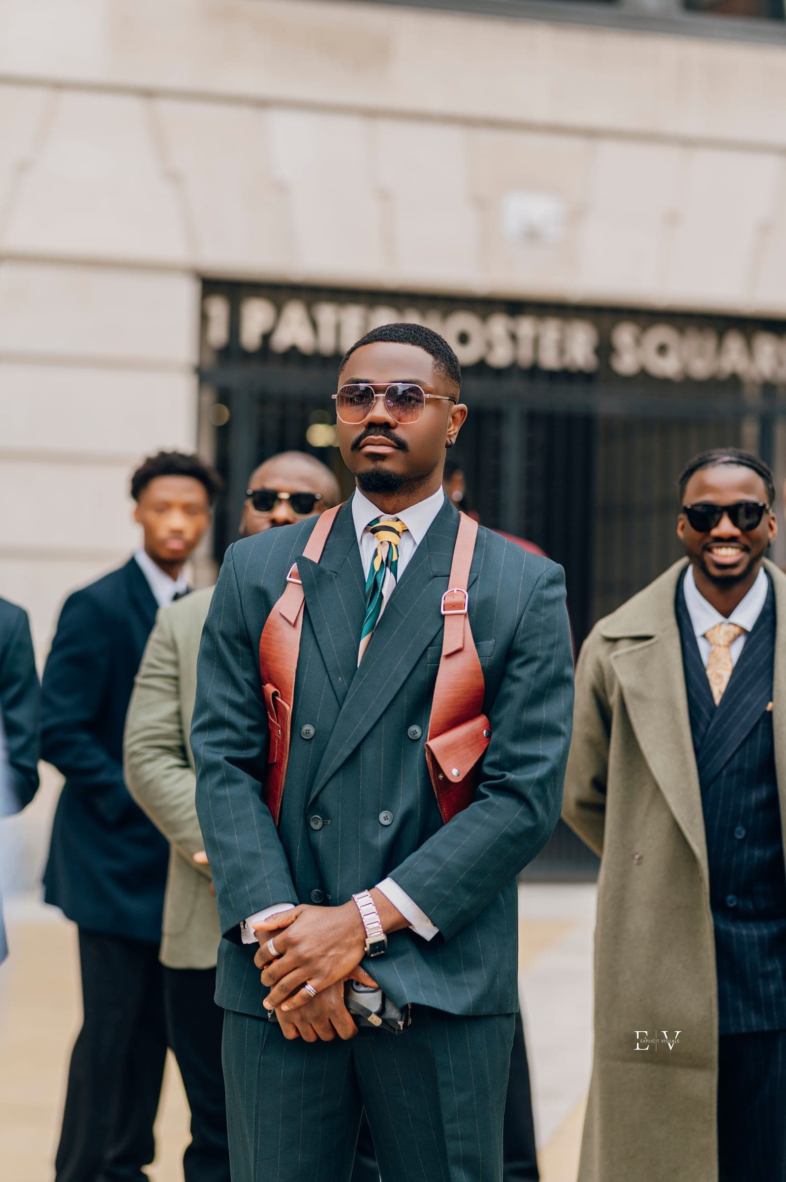 A man in a dark green double-breasted suit and a tan leather harness bag, hands folded in front of him, a Paternoster Square sign and other guests out of focus behind.