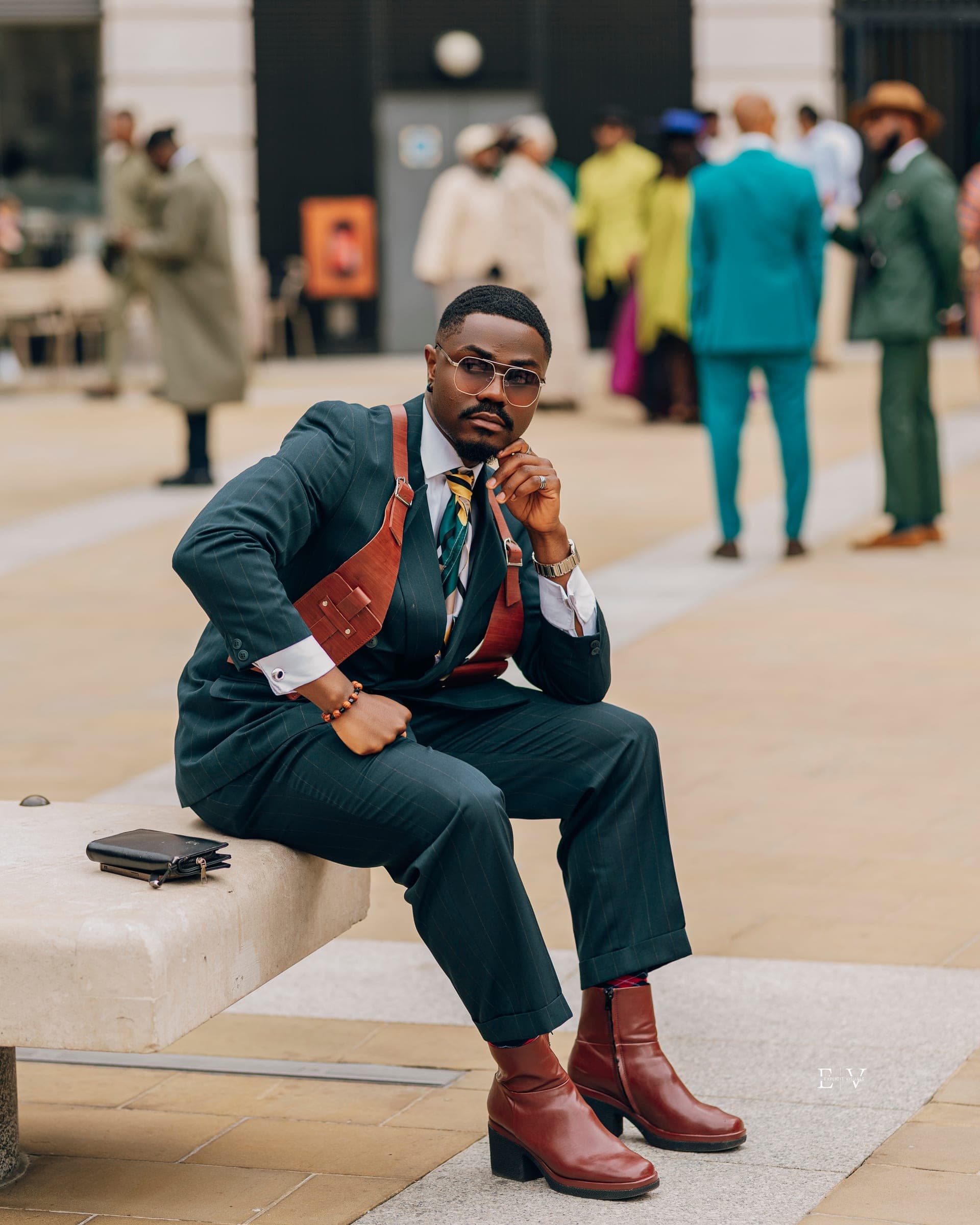 The same man seated on a stone bench with one hand to his chin, burgundy heeled boots crossed on the paving and a wallet set down beside him.