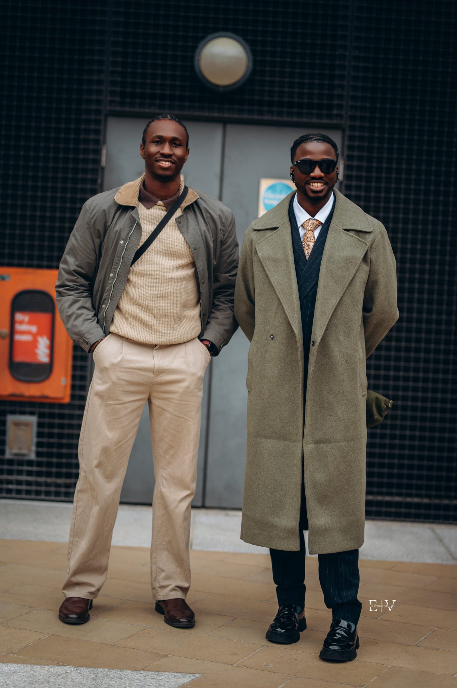 Two men in front of a dark mesh wall — one in an olive bomber over a cream knit and cream trousers, the other in a long olive overcoat, shirt and tie.