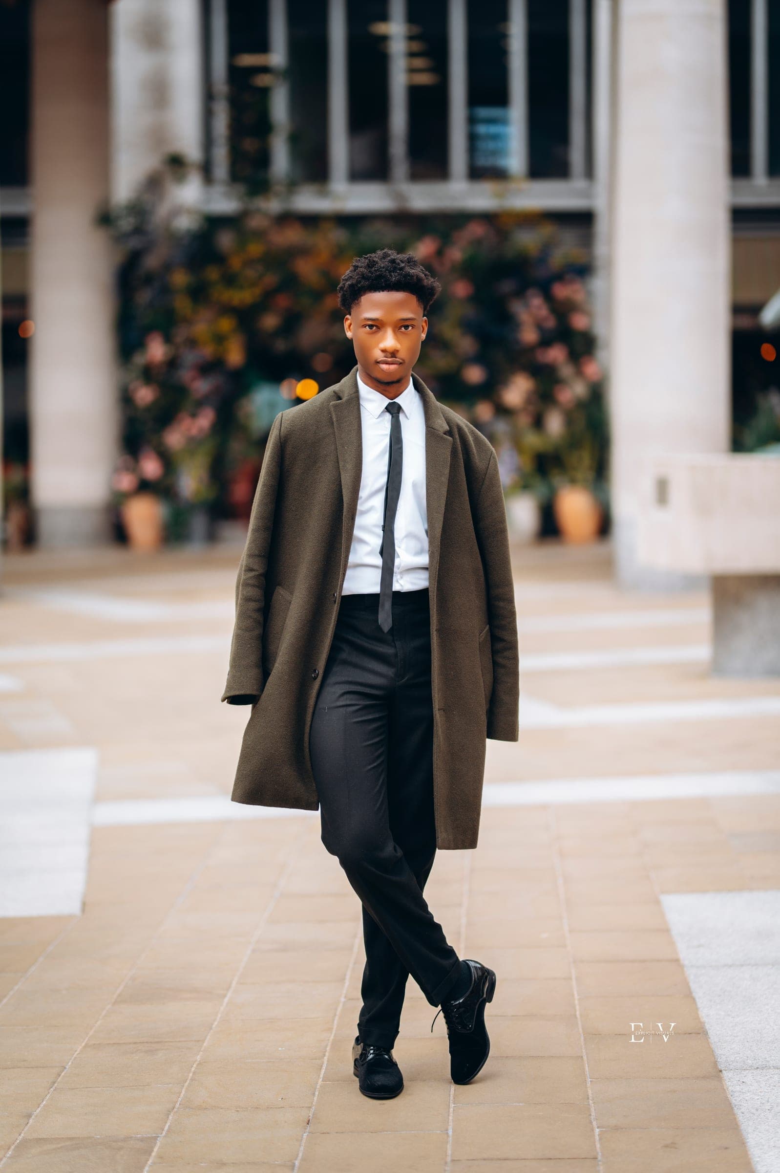 A young man walking toward the camera in an olive overcoat over a white shirt and black tie, flowers and a stone colonnade behind him.