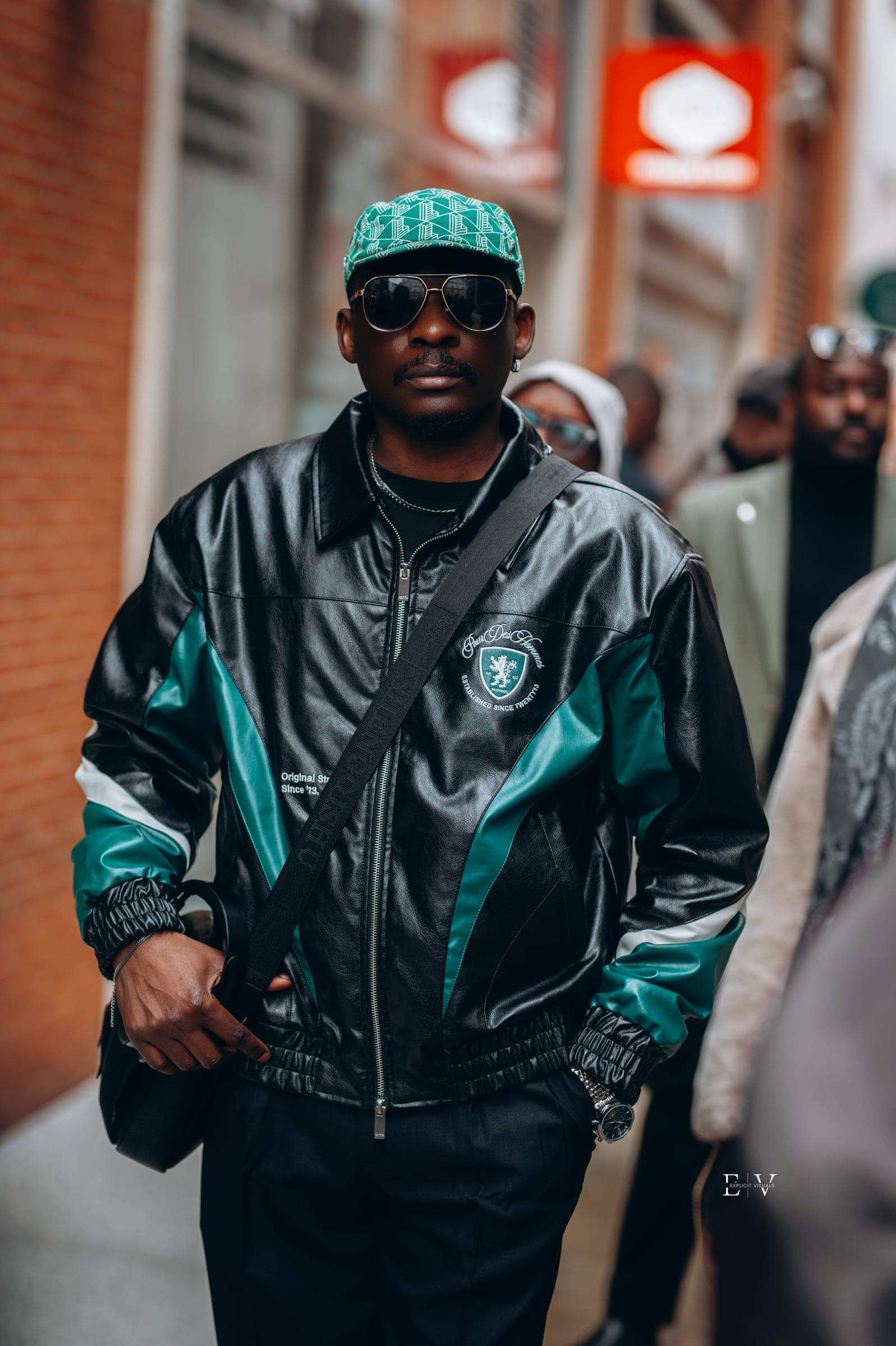 A man in a black and teal panelled leather bomber with a matching knitted cap and a crossbody strap, standing in a busy street.
