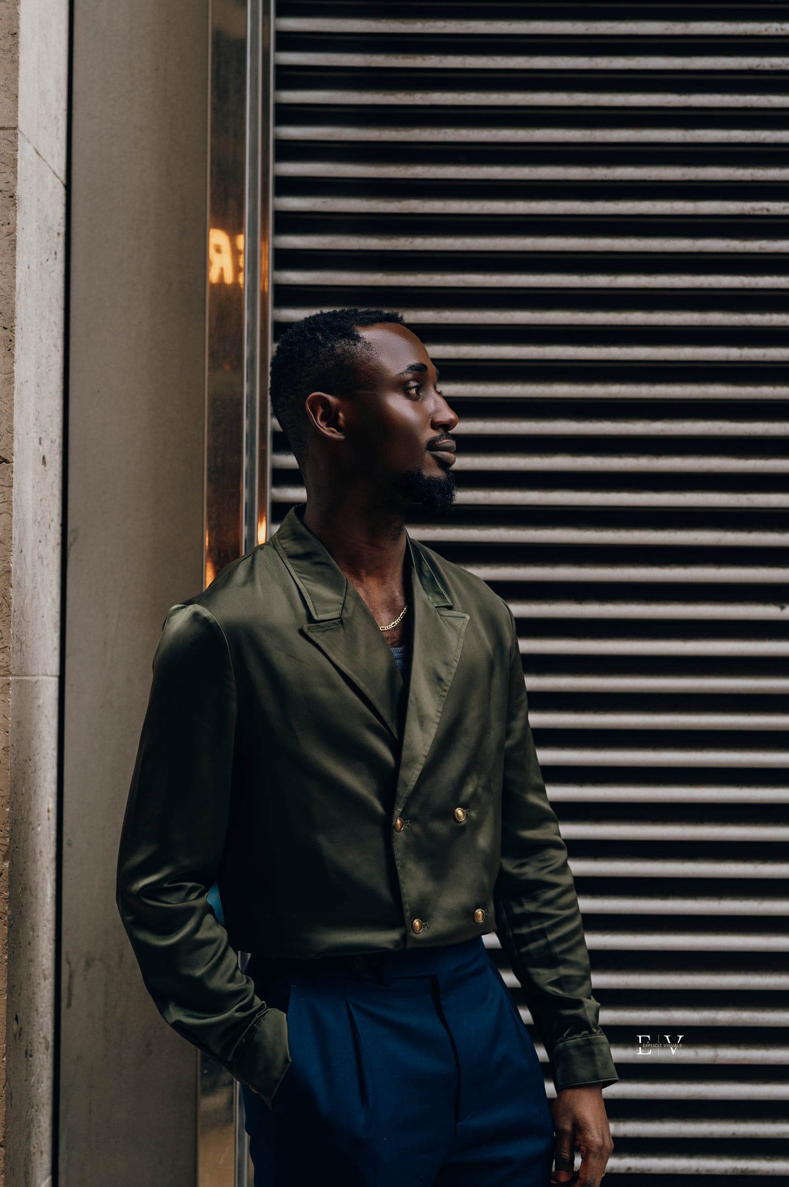 A man in an olive leather jacket and navy trousers leaning against a doorway beside a metal shutter, looking off to one side.