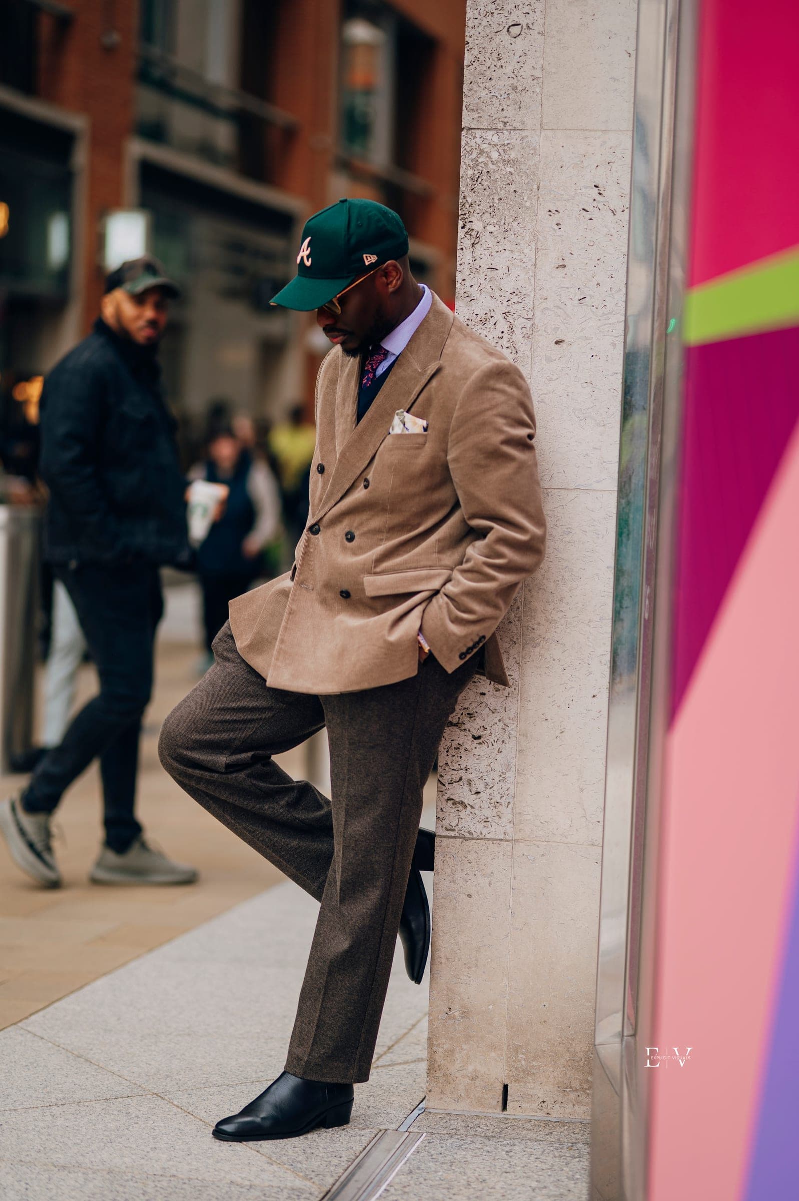 The same look full length, one foot up against a stone corner, head down, a bright painted panel to his right.