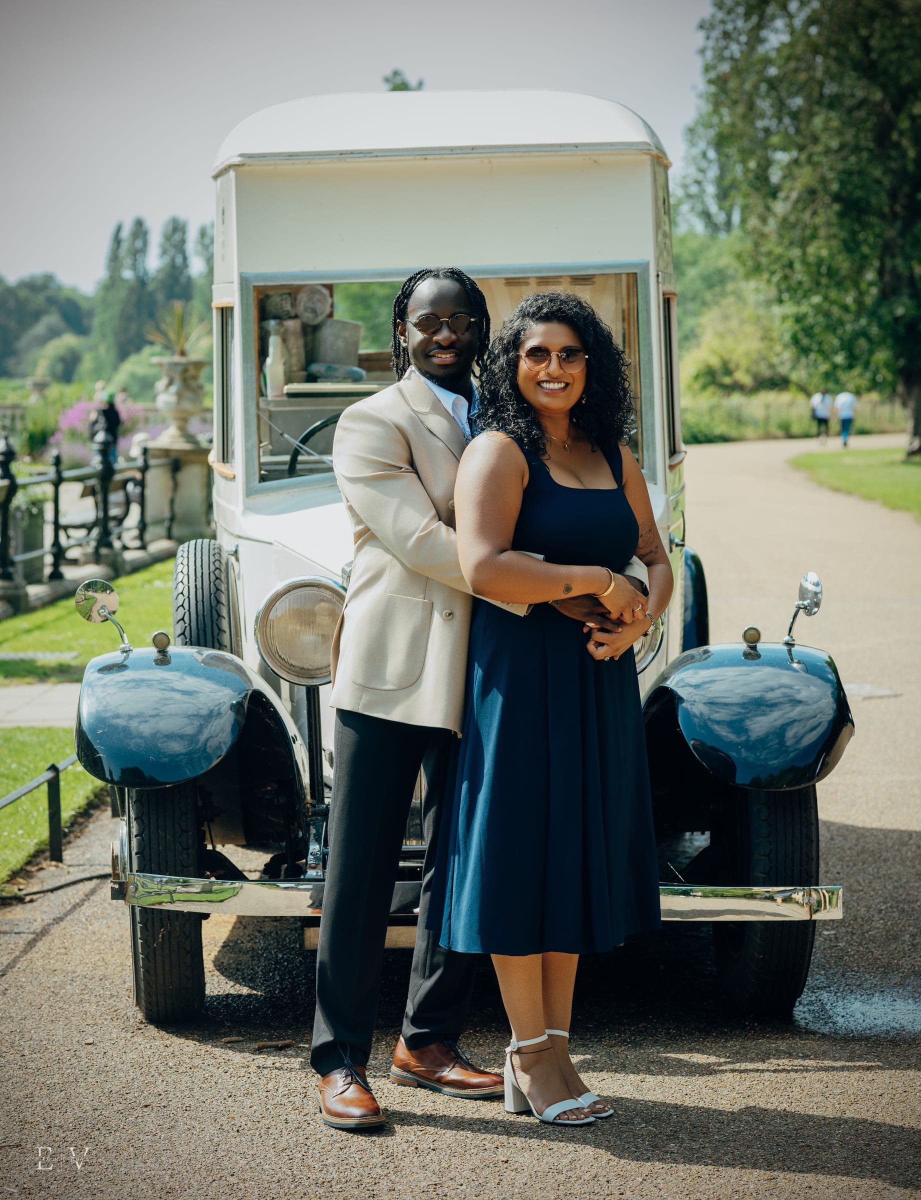 The couple hold each other in front of a vintage cream ice-cream van parked on a garden path, looking straight to camera.