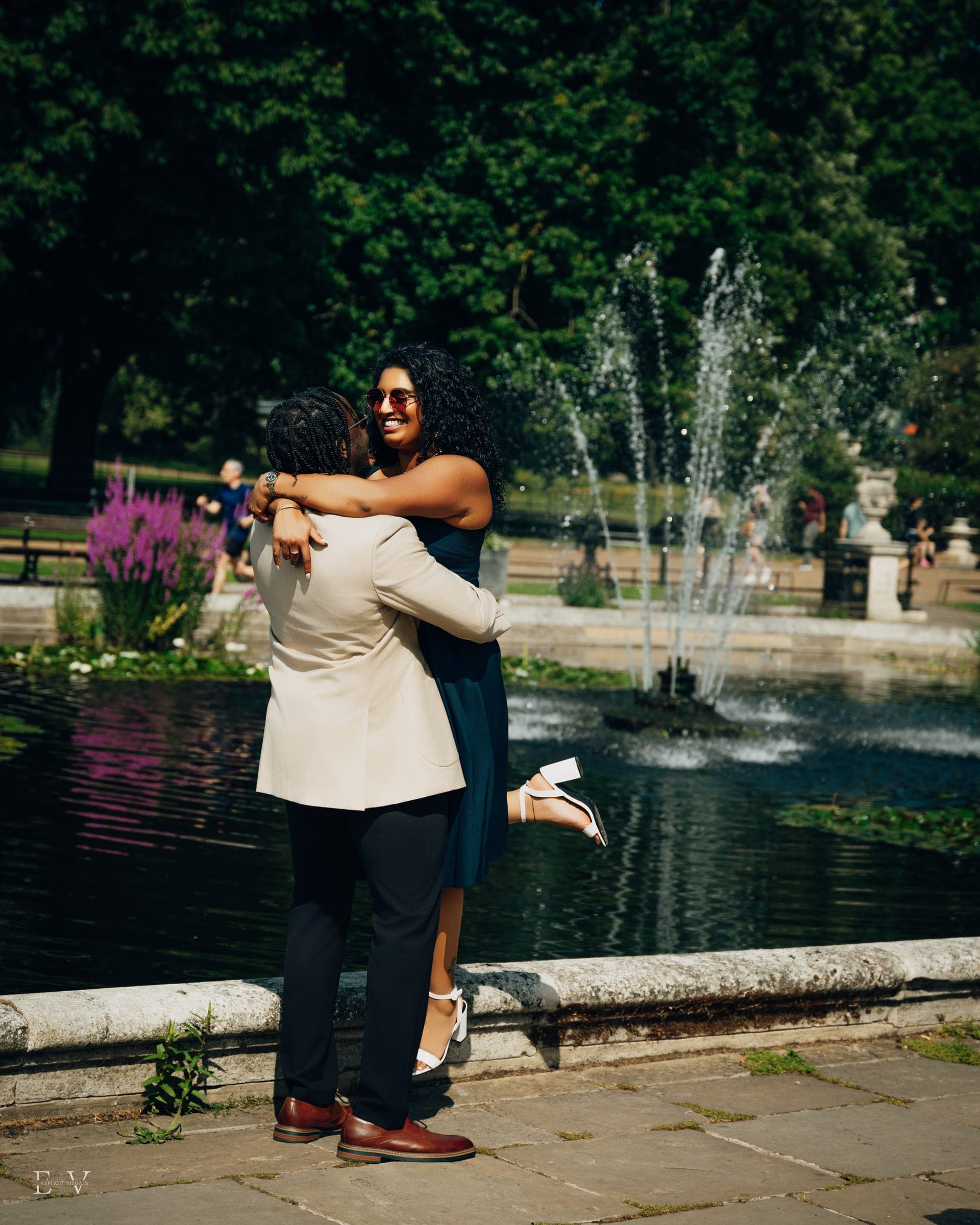 A laughing embrace at the edge of an ornamental pool, one lifted clear off the ground, fountain jets rising behind them.
