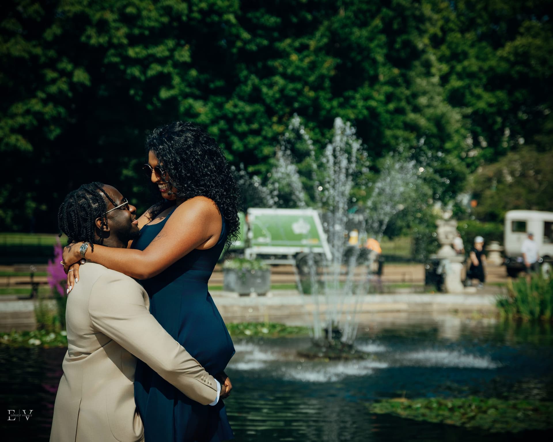 Held close at the pool's edge, one resting a head against the other's shoulder, a fountain catching the light behind.