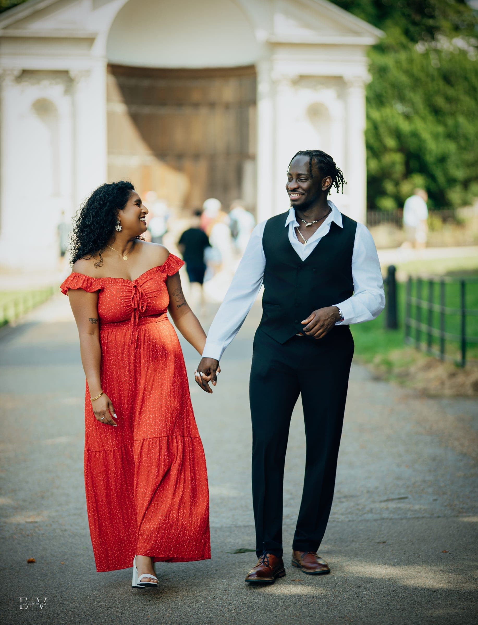 Walking hand in hand toward the camera in the red dress and black waistcoat, a white stone garden gateway behind them.