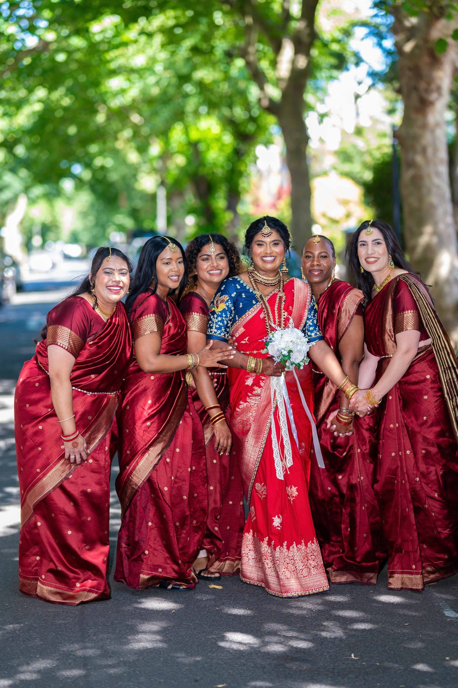 The bride and her five bridesmaids pressed together on a plane-tree avenue, hands linked, sunlight coming through the canopy.