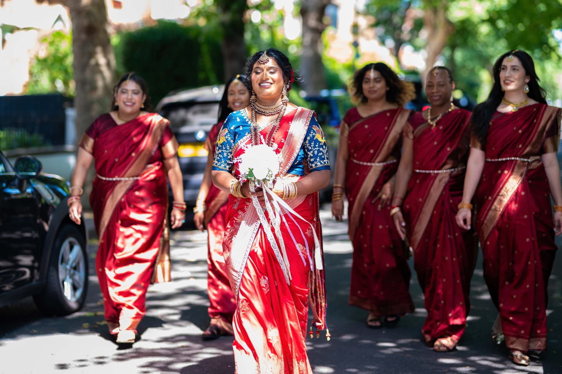 The bride walks up the road toward the camera with her bouquet, bridesmaids fanned out behind her.