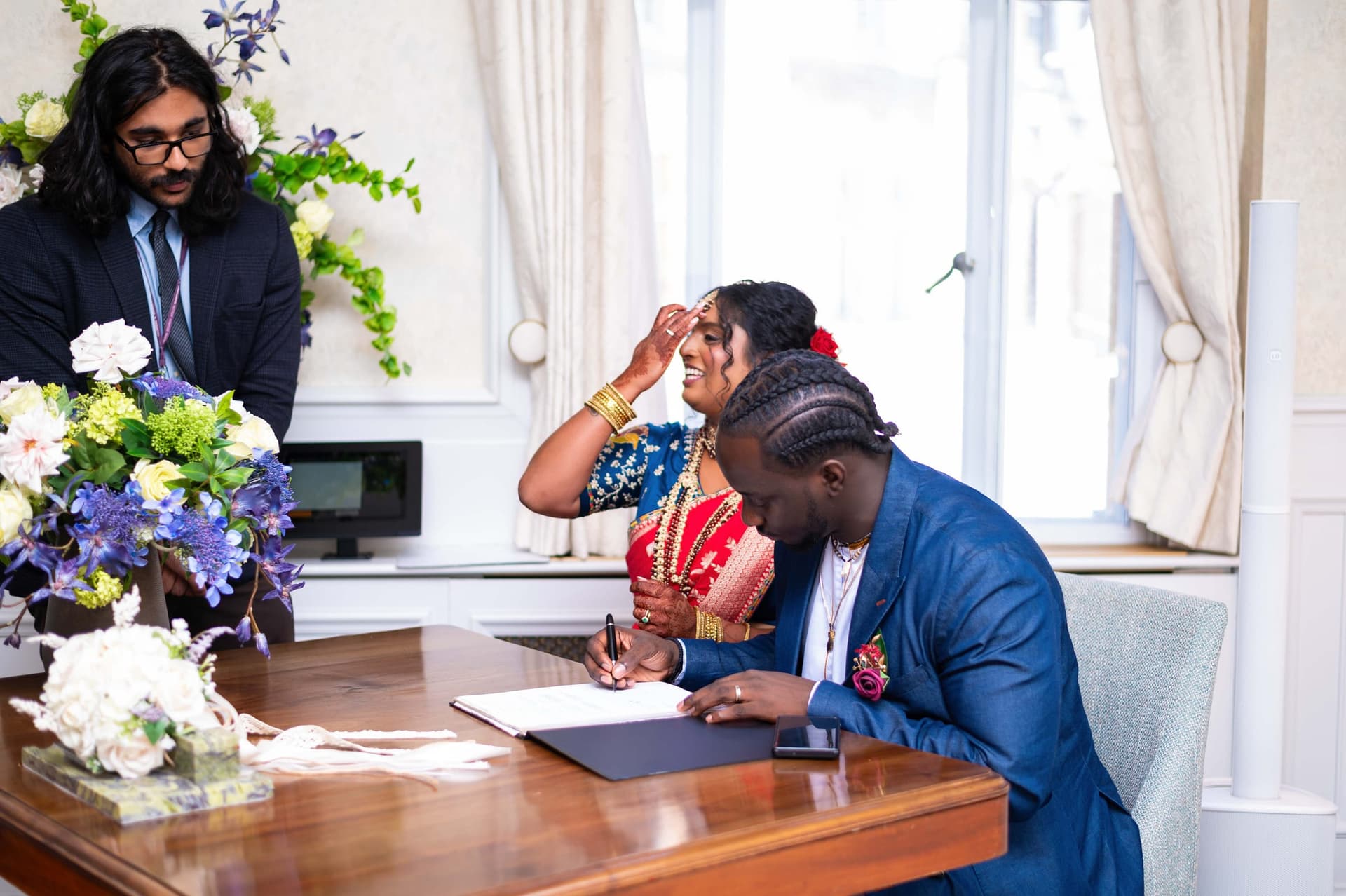 The groom signs the register at a wooden table; behind him the bride laughs with a hand to her forehead.