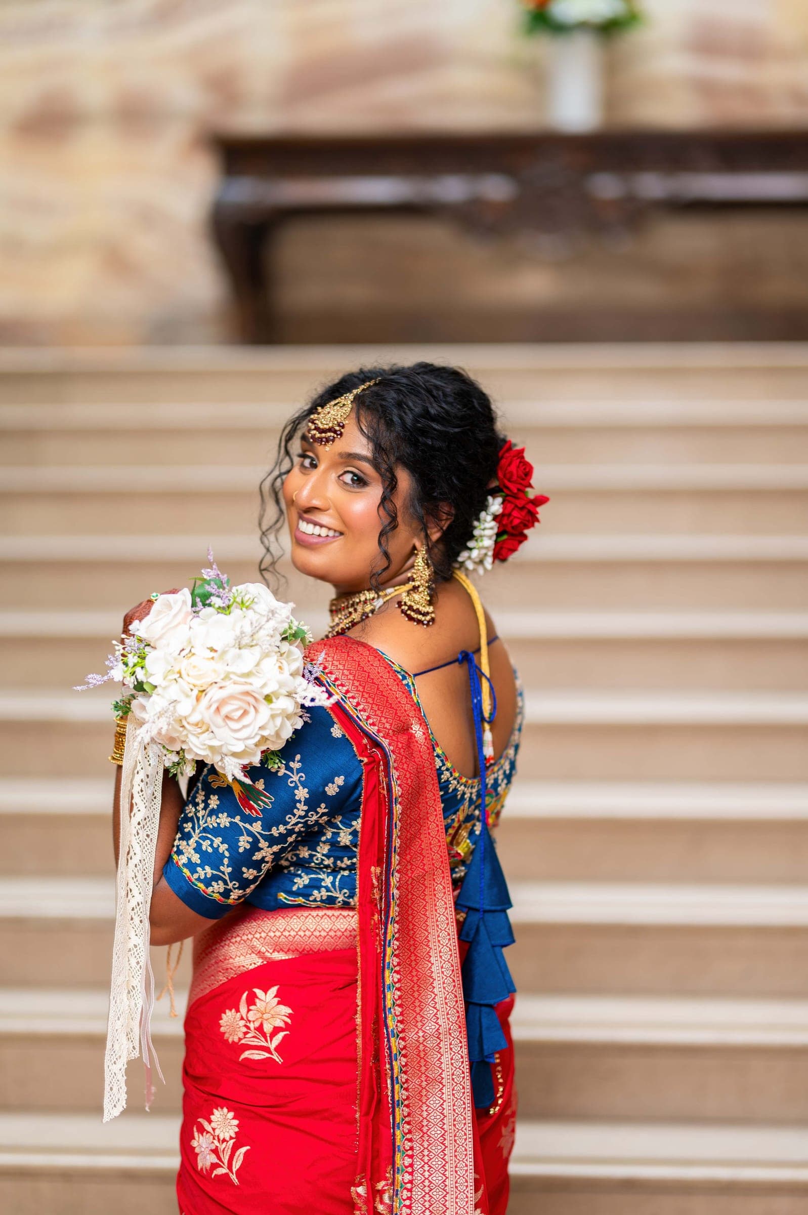 The bride turns back over her shoulder on the stairs with her bouquet raised, a red rose and jasmine pinned in her hair.