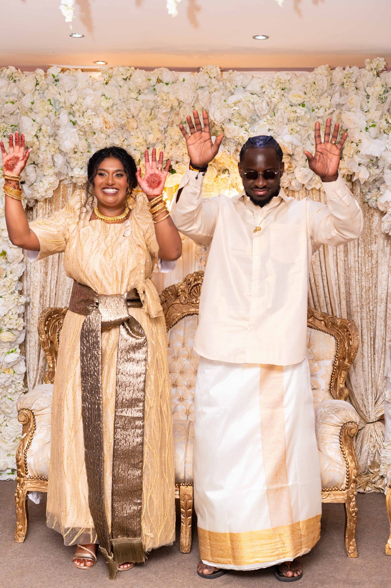 At the reception, the couple stand in cream and gold before a wall of white flowers with both hands raised to show their henna.