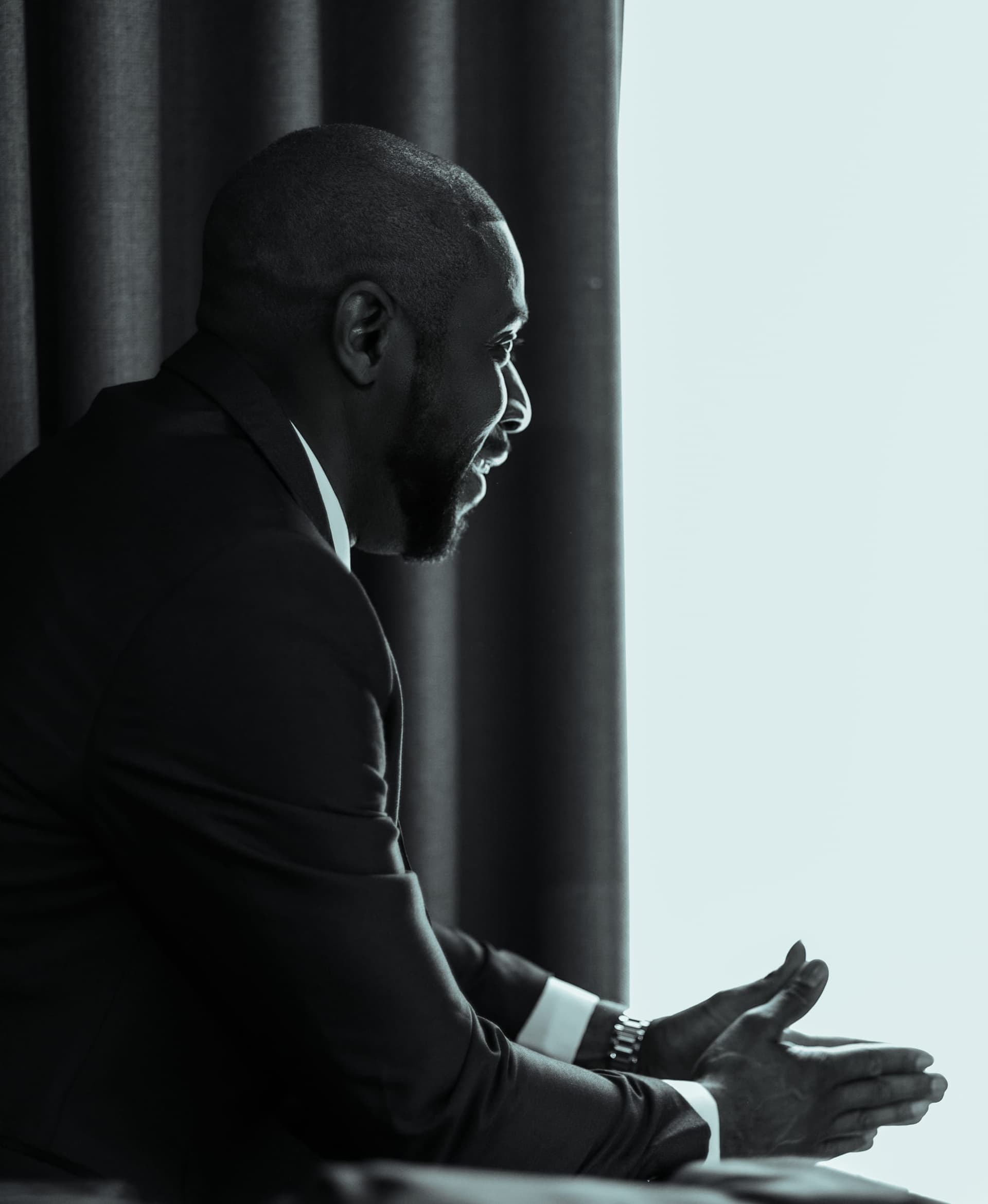 In black and white, the groom sits forward by a curtained window, hands loosely clasped, waiting.