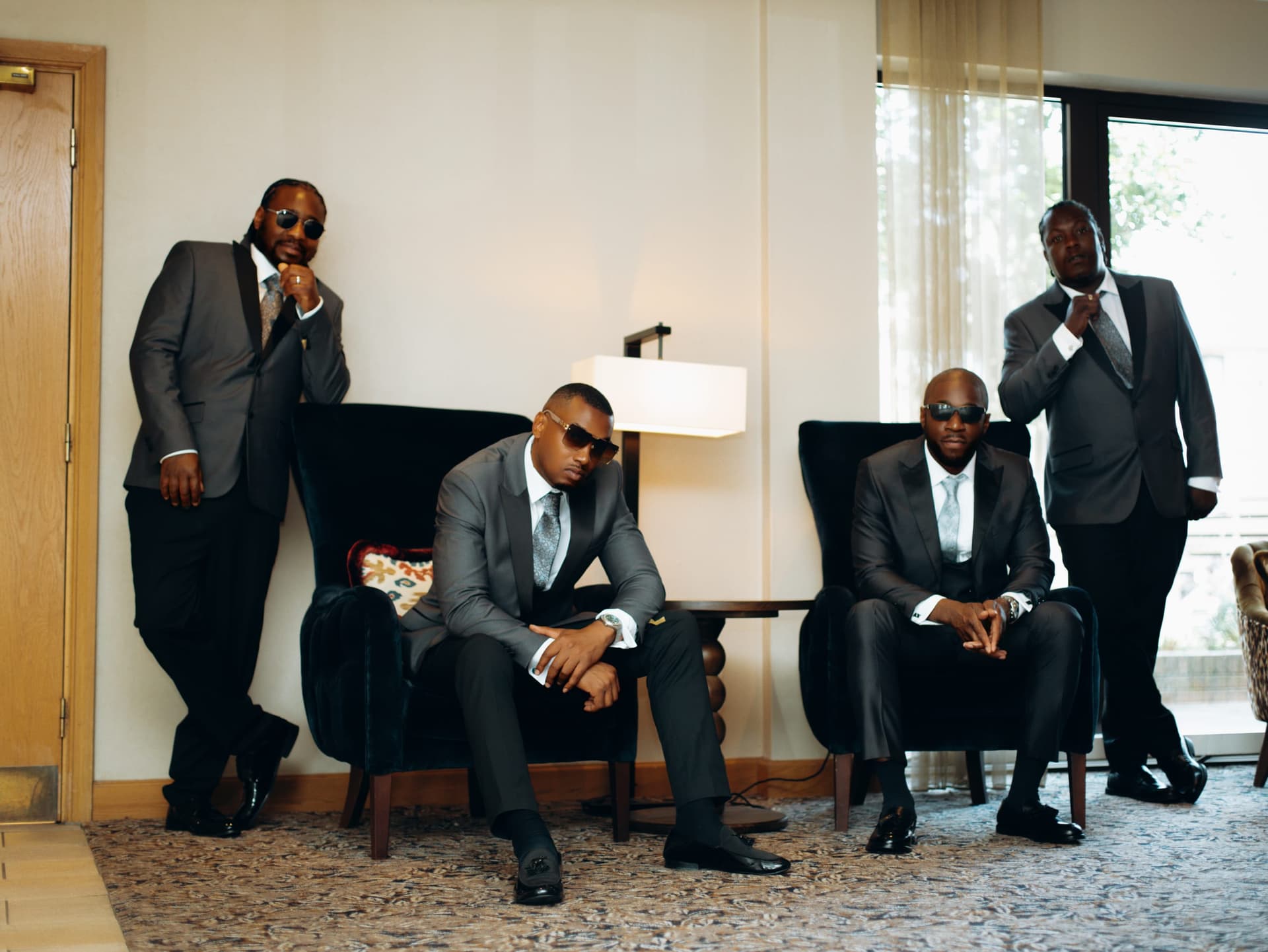Four groomsmen in grey and charcoal suits and dark glasses arranged around the armchairs of a hotel suite.