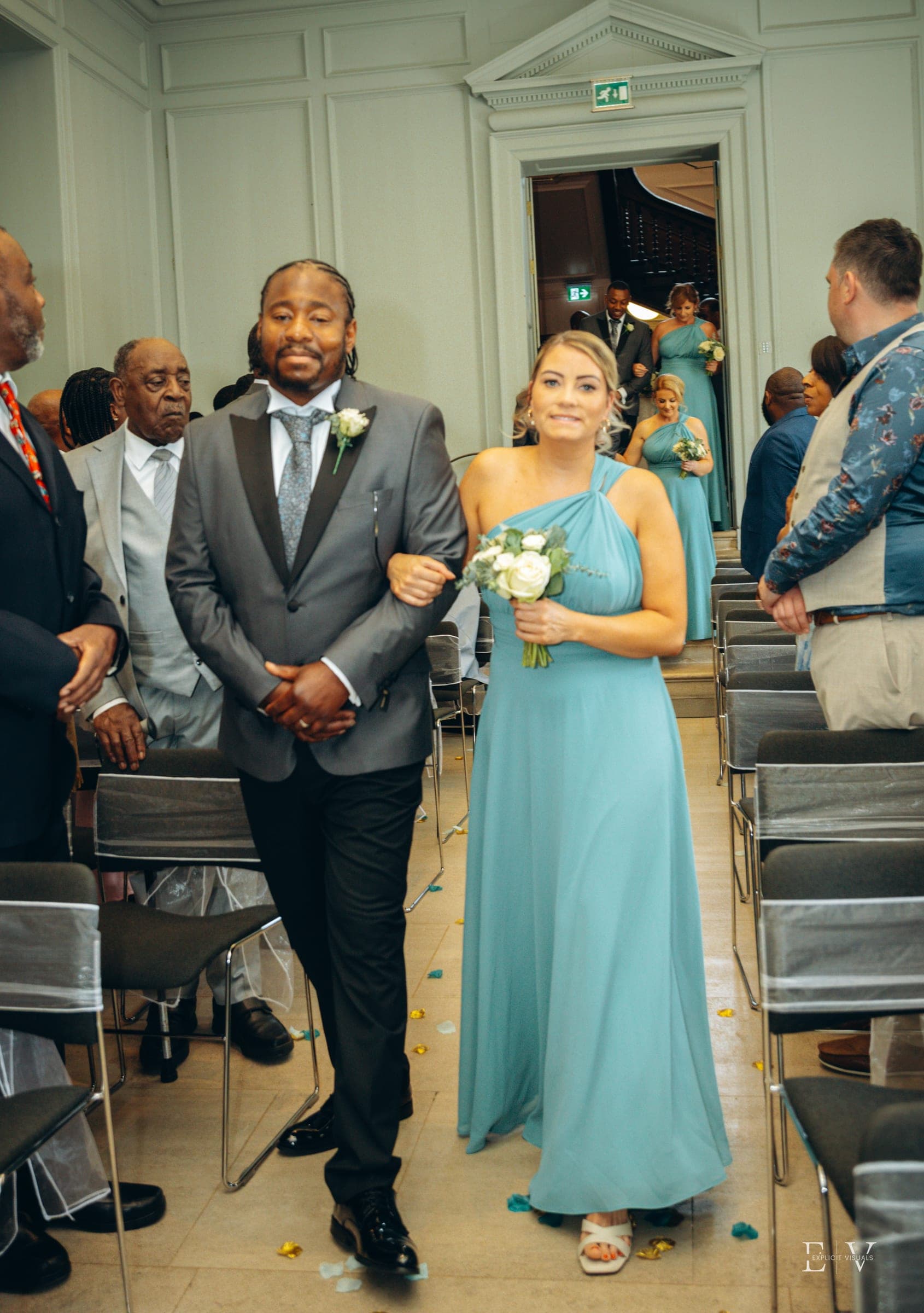 A bridesmaid in a teal one-shoulder gown is walked down the aisle on the arm of one of the party, guests standing on both sides.
