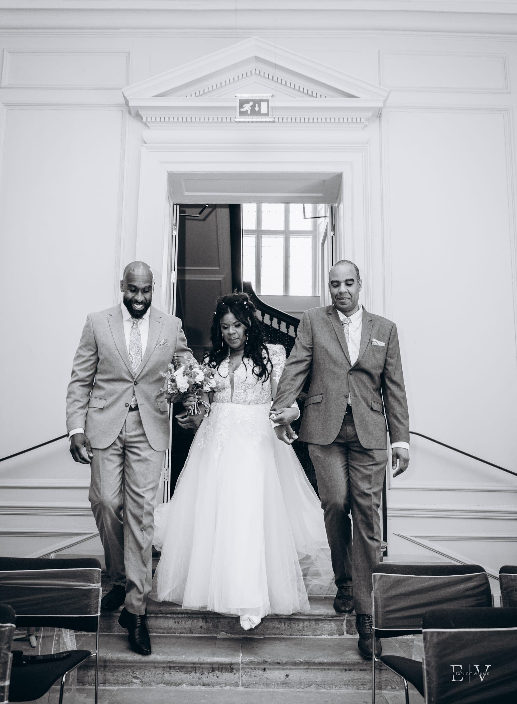 In black and white, the bride comes down the steps into the ceremony room flanked by two men in light grey suits.