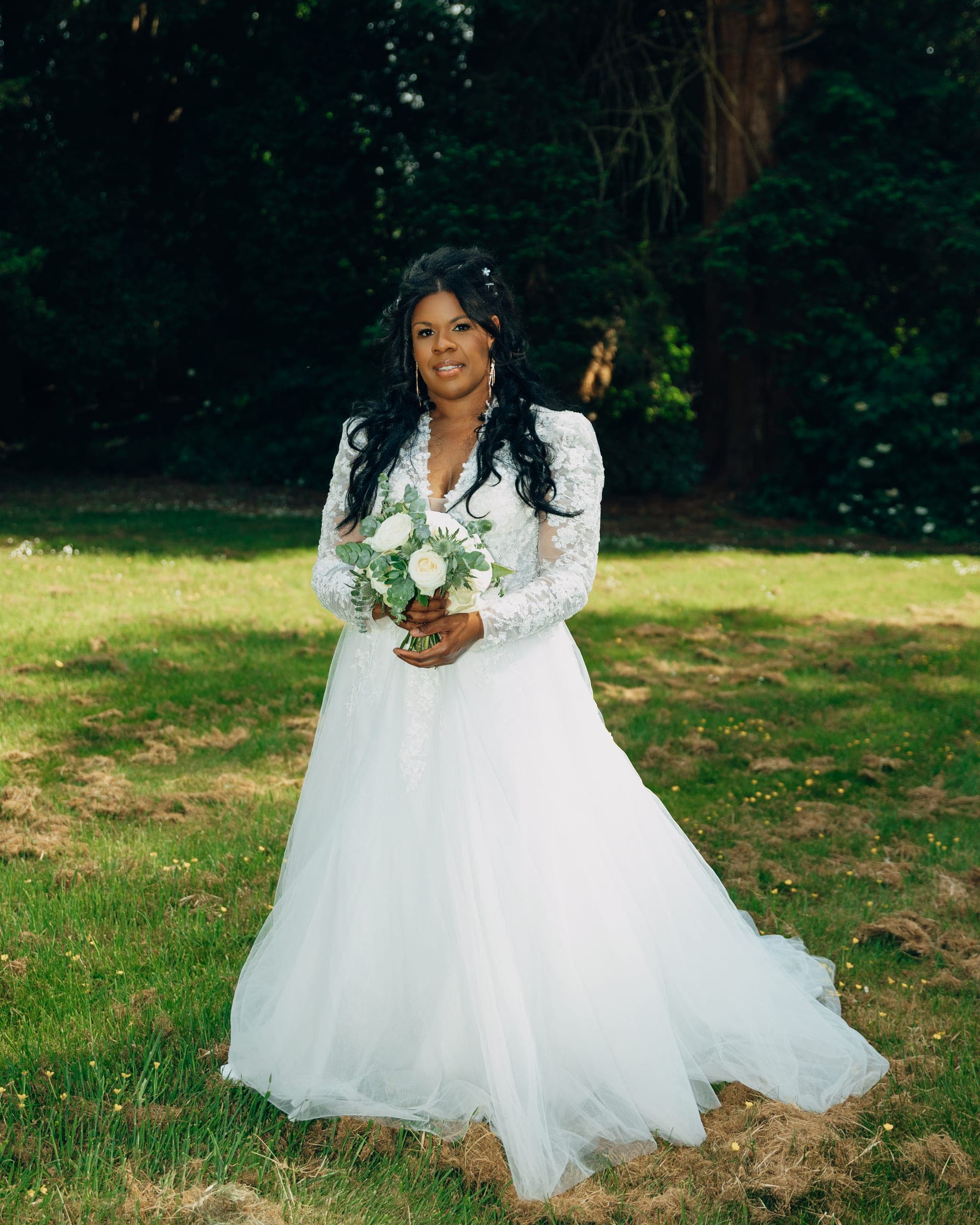 A full-length portrait of the bride on the lawn with her white rose bouquet, tall dark trees behind her.