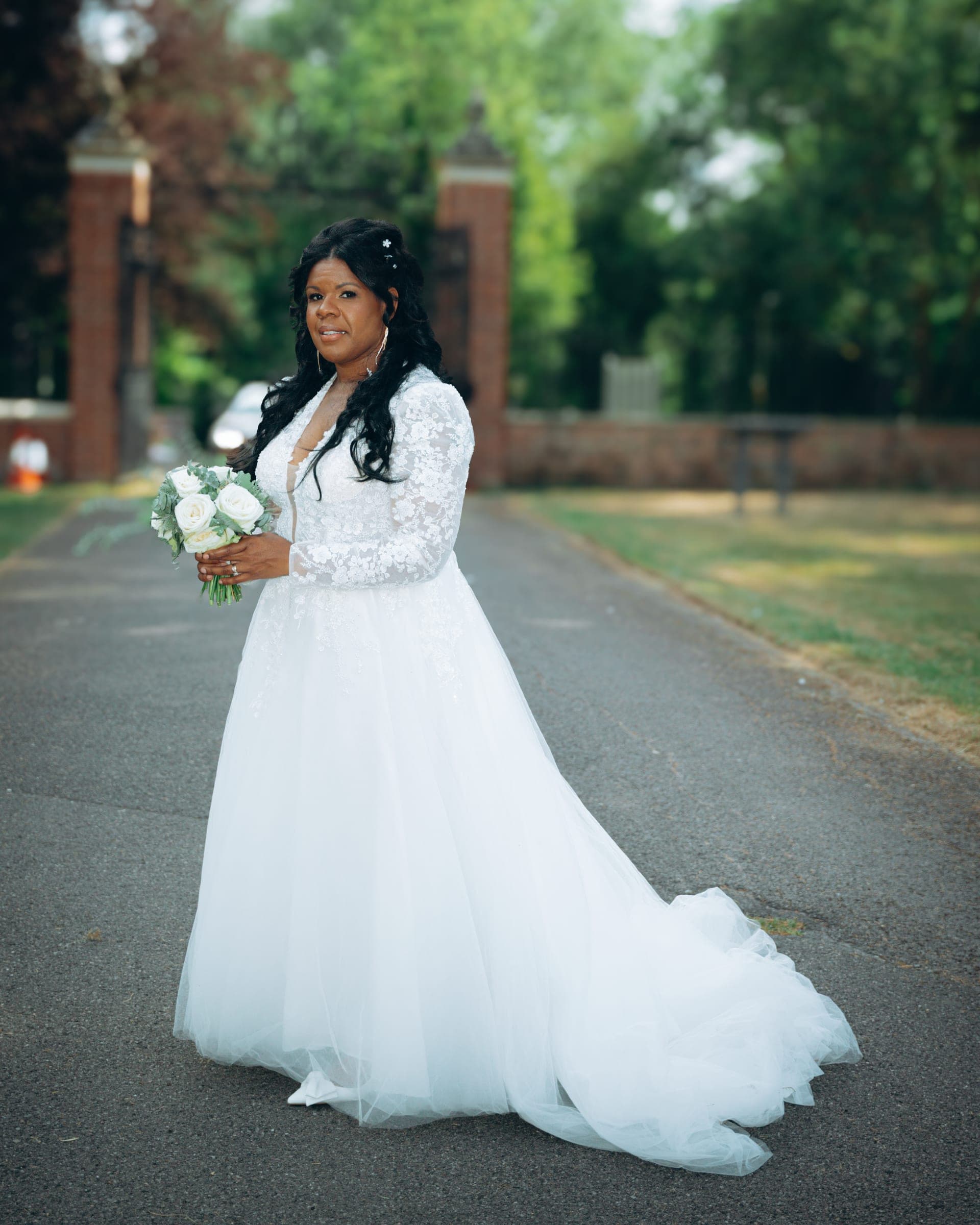 The bride standing on the drive by the brick gateposts, train trailing behind her on the tarmac.