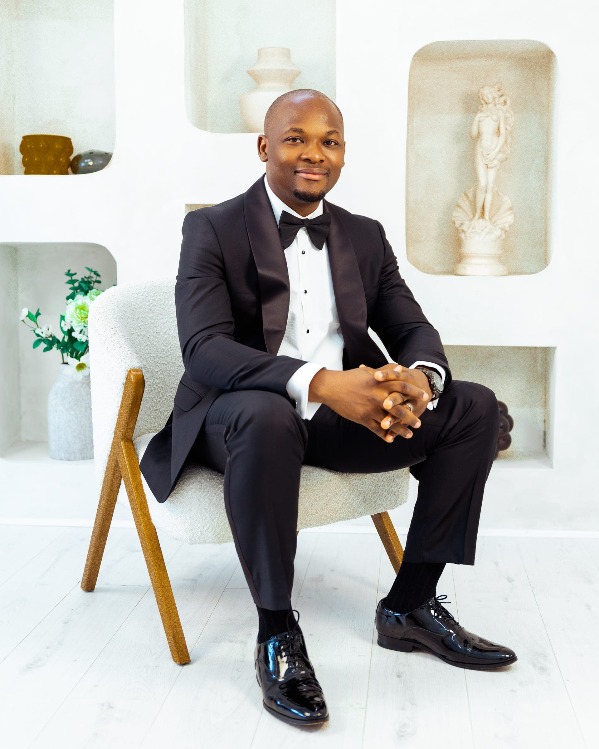 Seated in a cream bouclé chair with his fingers laced, patent shoes crossed at the ankle, sculpture and vases set into the alcoves either side.