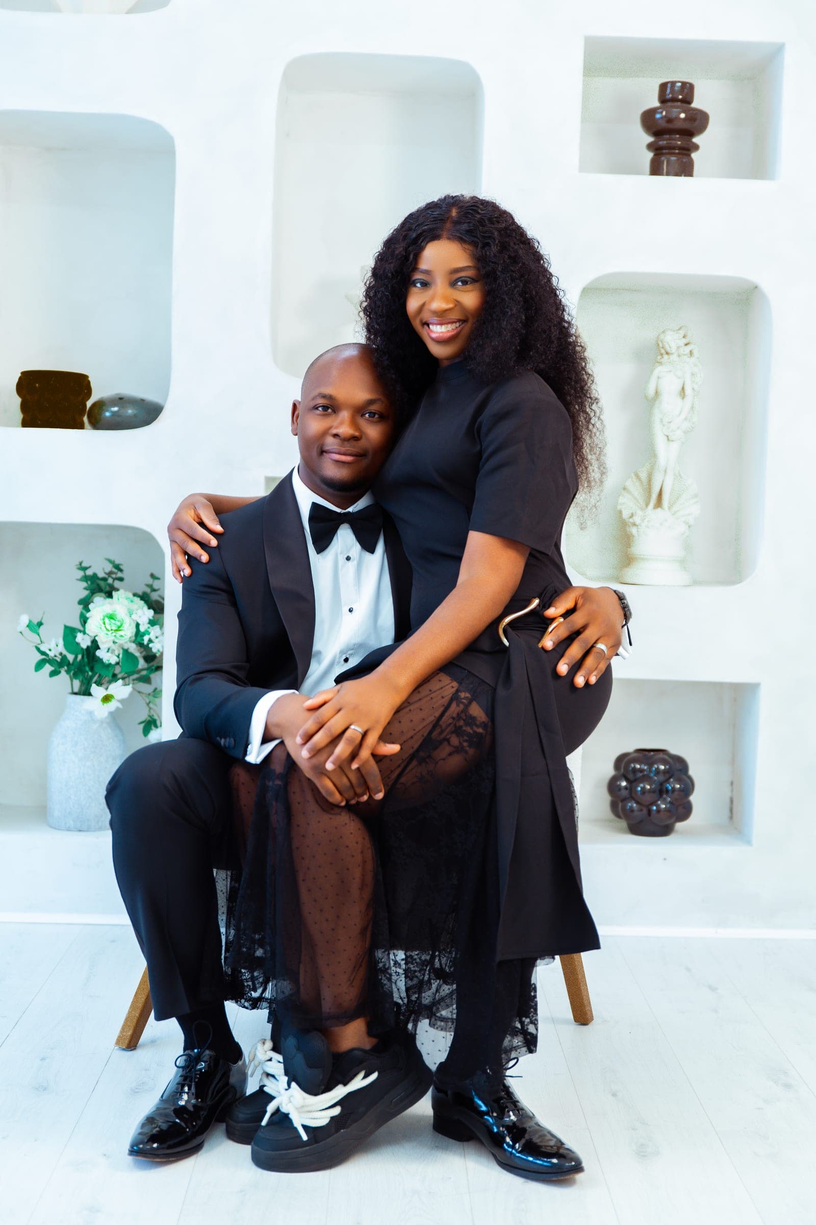 His wife perches on his knee in a black dress and sheer skirt, one arm around his shoulders, both smiling to camera.