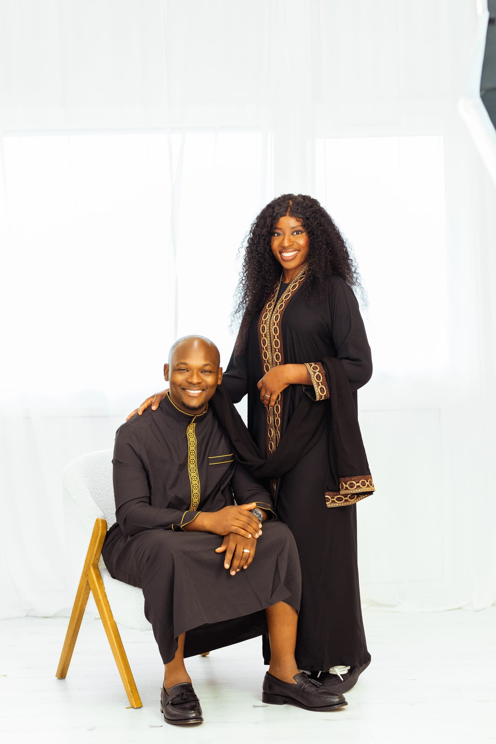A third look — a black kaftan with gold embroidery, seated, his wife standing beside him in a matching black and gold abaya.