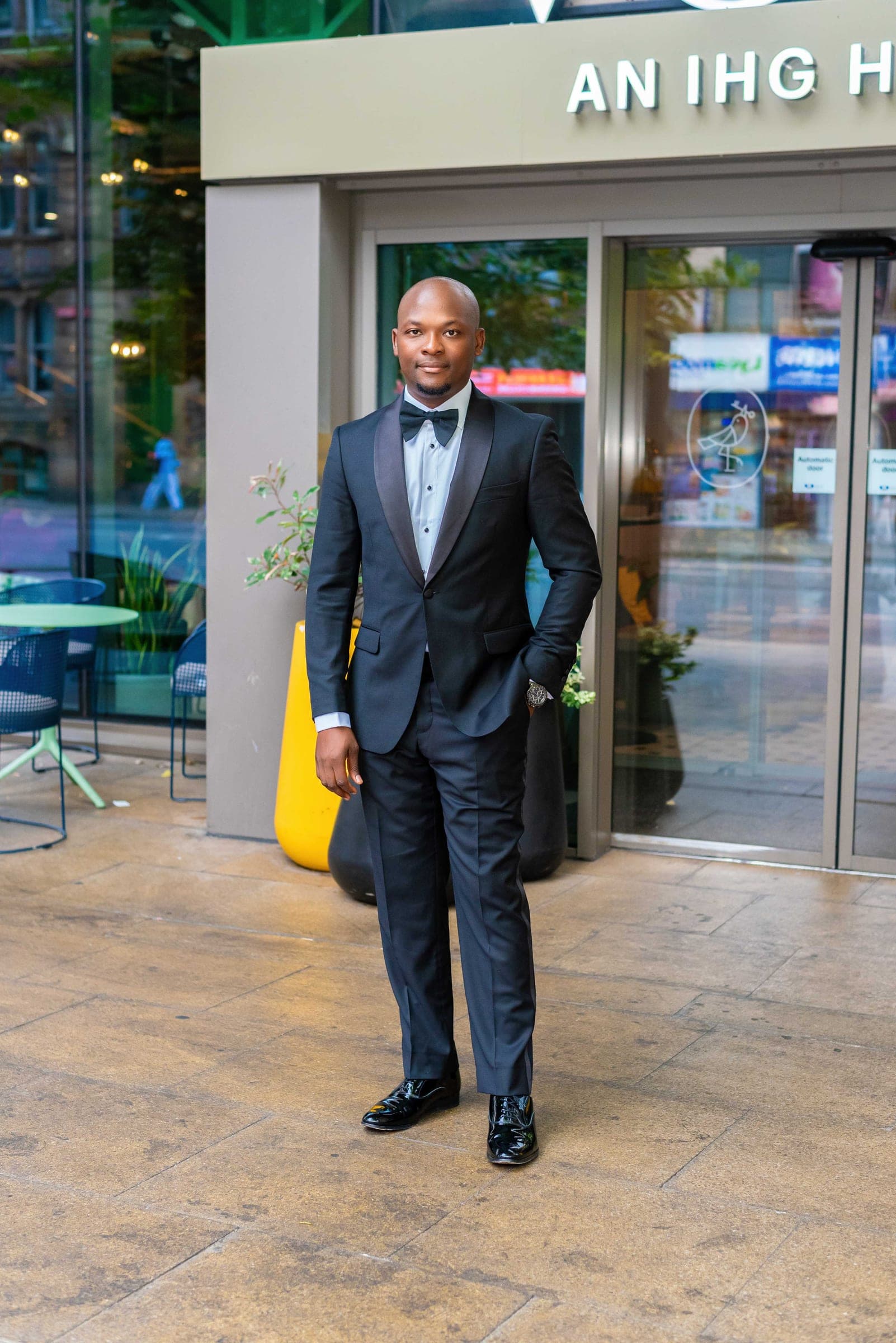 The same spot framed tighter — hands in his pockets, bow tie and patterned shirt front, the street reflected in the glass.