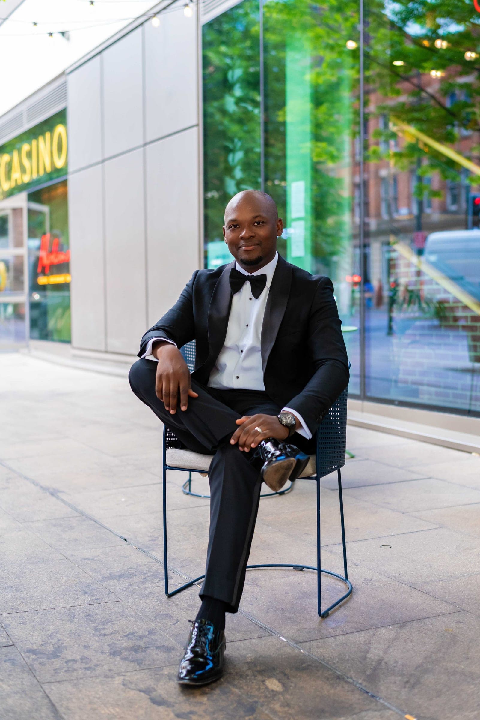 Seated on a low metal stool on the pavement, one ankle across his knee, a lit shopfront and passing traffic behind.