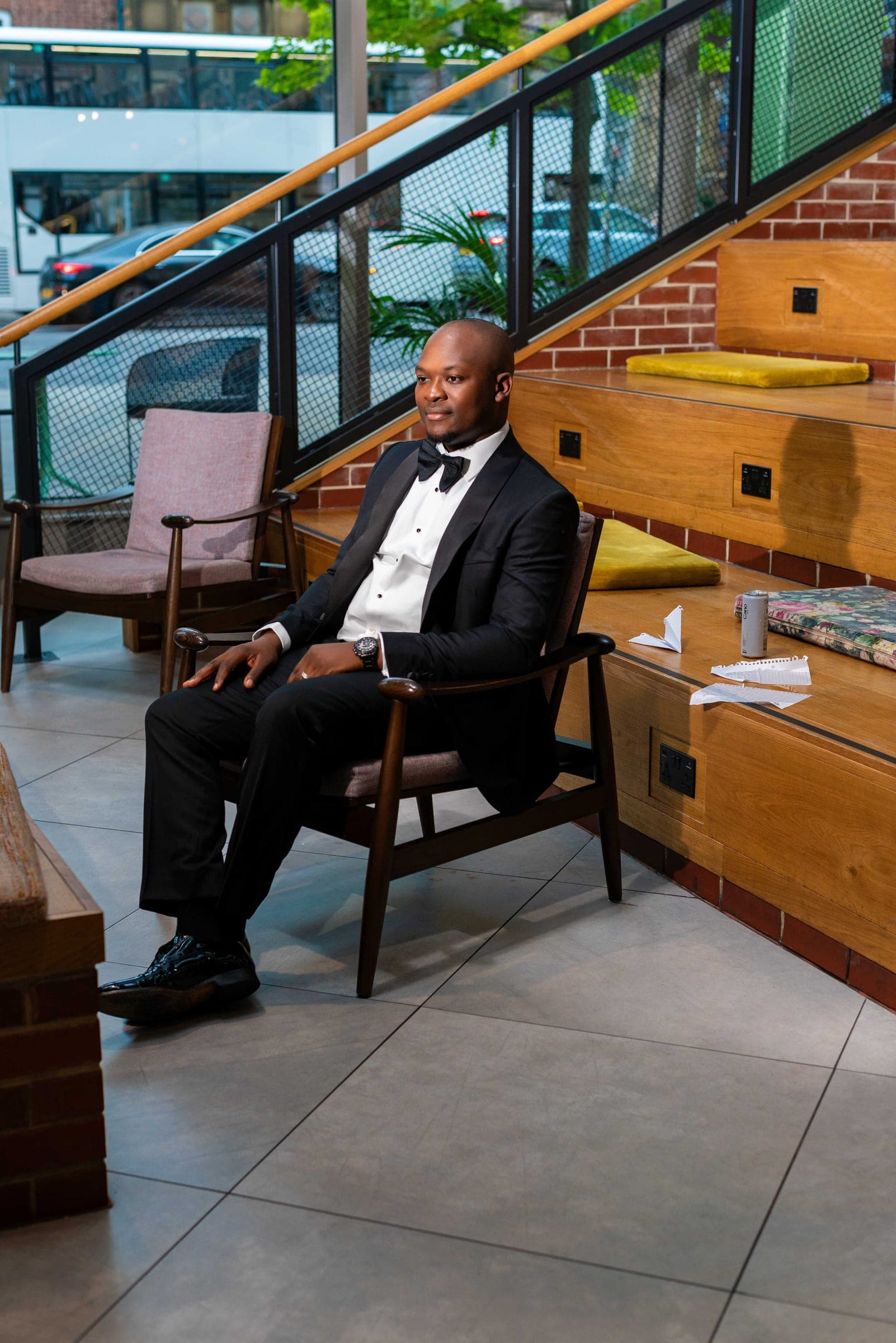 Sitting back in a mid-century armchair in the hotel lobby, a staircase and brick banquette seating behind him.