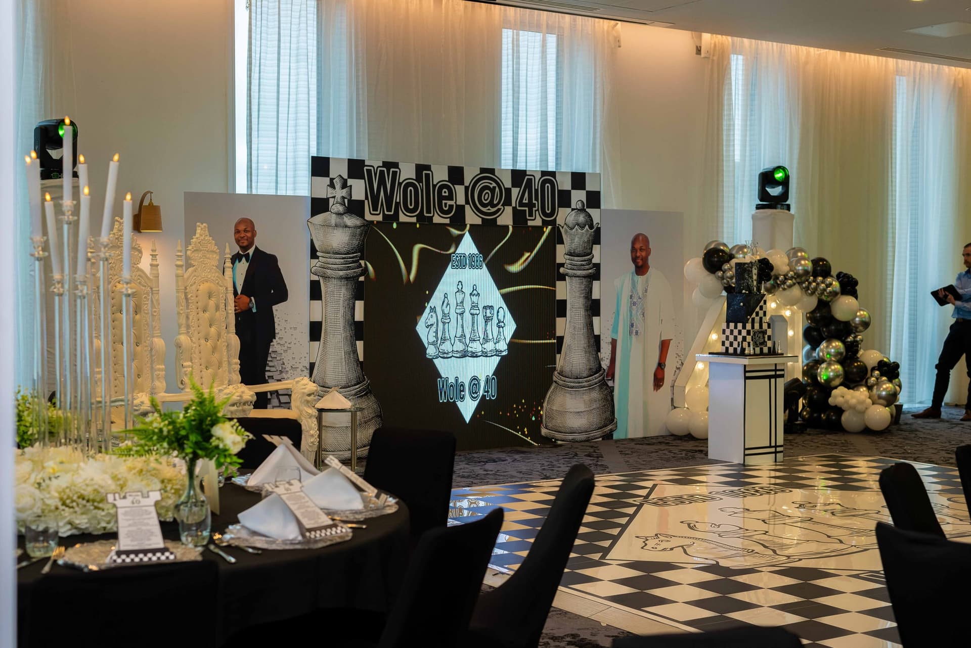 The room set and empty before the doors open — black-clothed tables, silver candelabra, a chequered dance floor and a balloon arch by the stage.