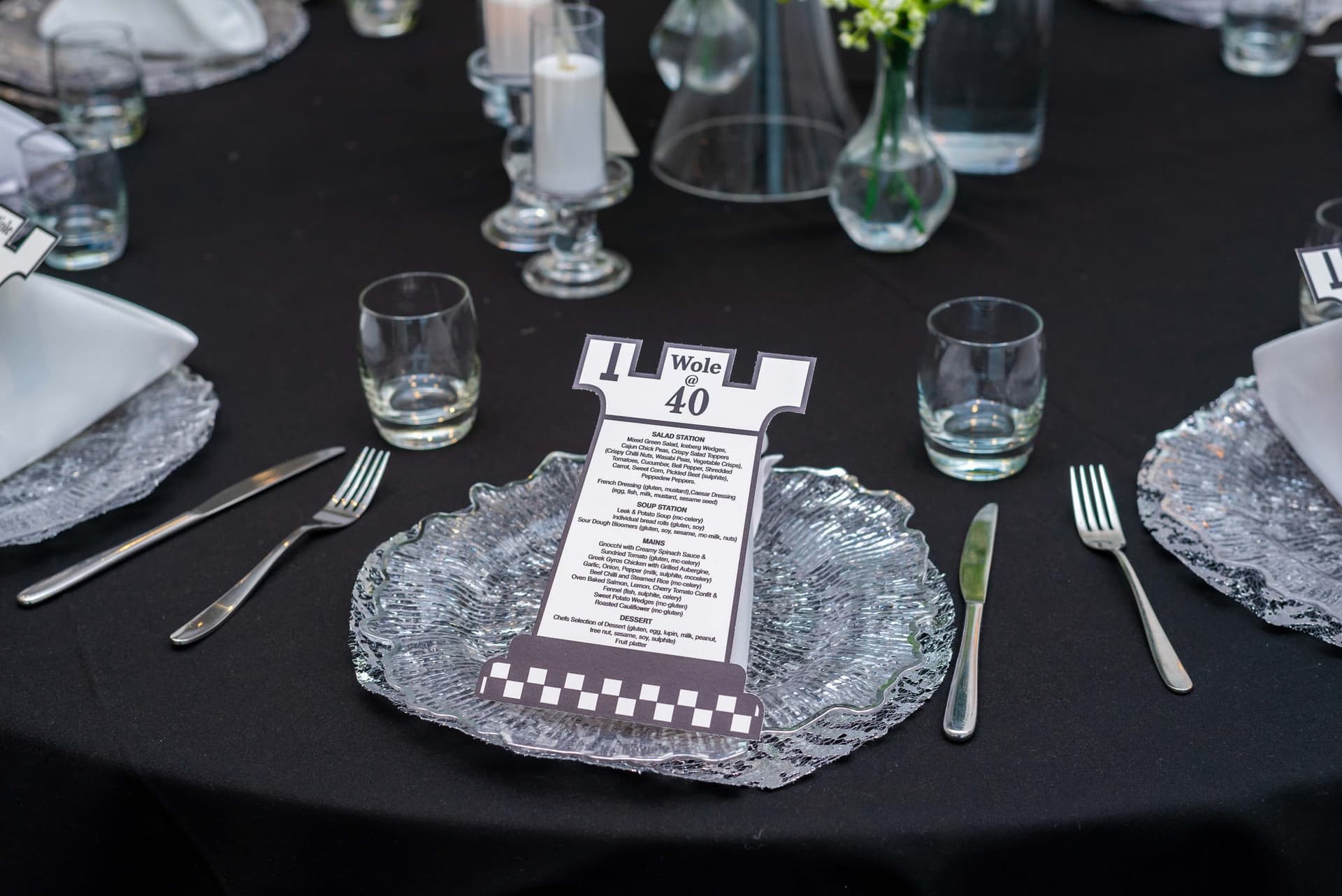 A place setting in close — a menu card cut into the shape of a chess rook, reading Wole at 40, on a textured glass charger between the cutlery.
