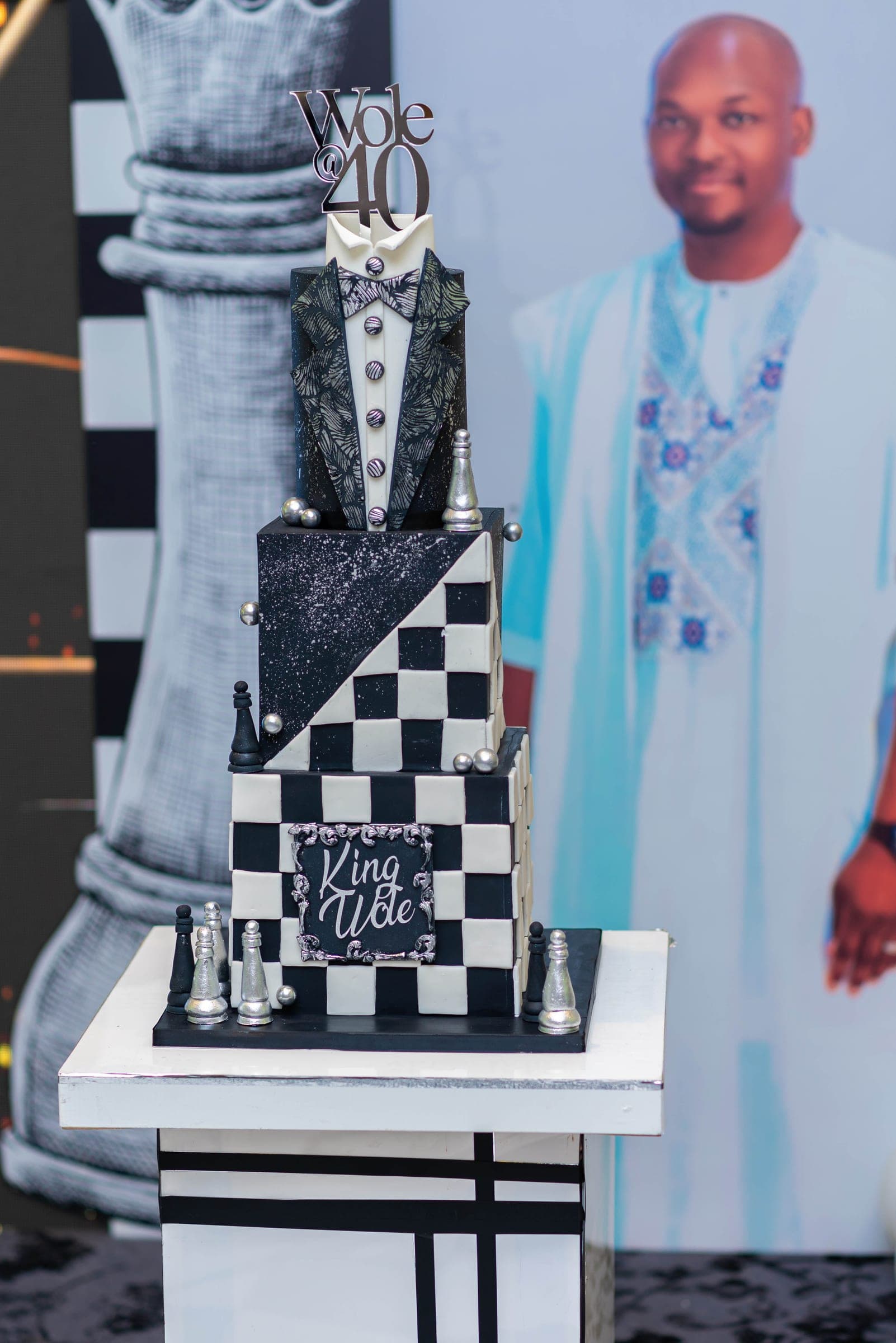 The same cake from the side, framed against a printed chess king and the life-size cutout in blue Nigerian dress.