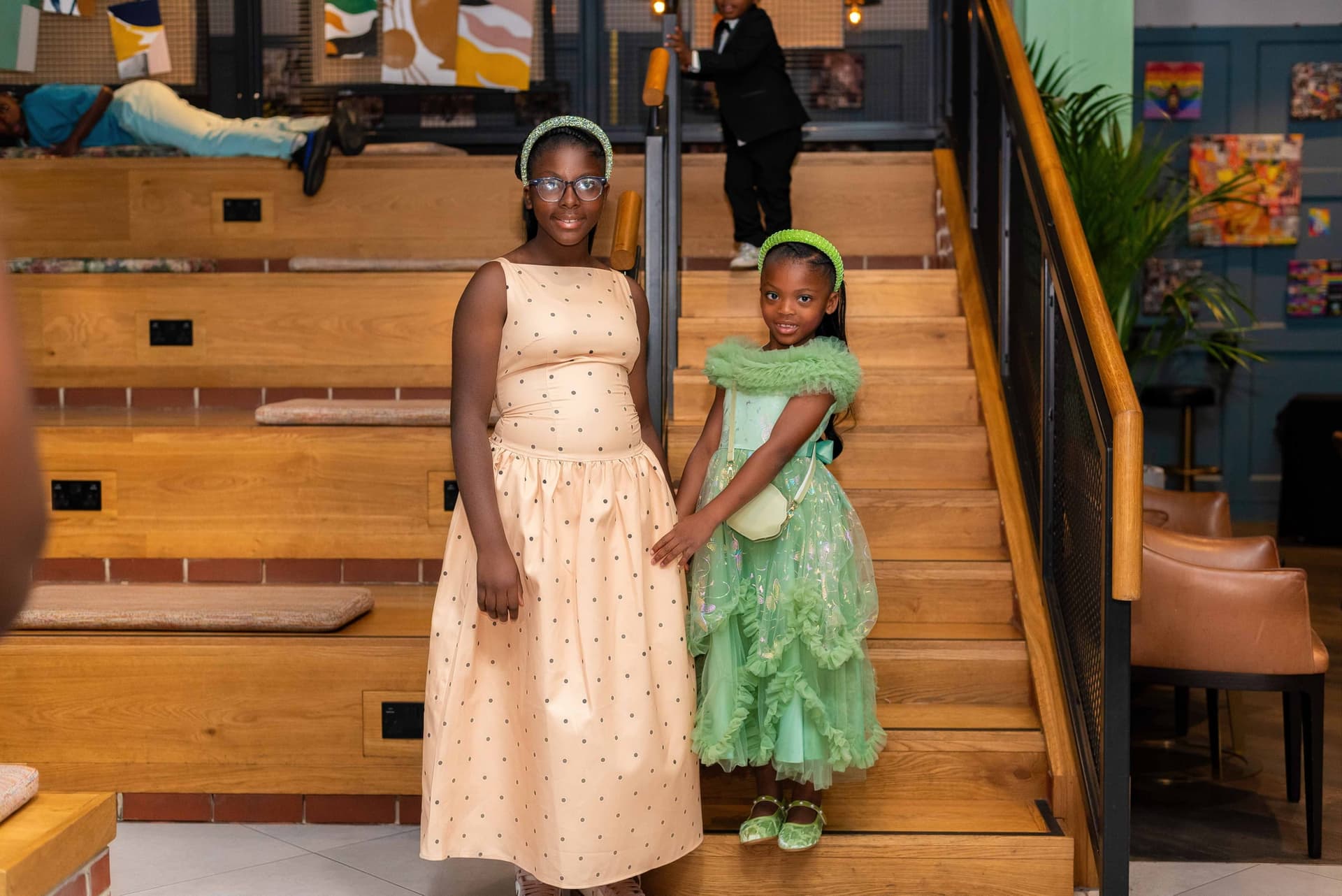 Two young guests on the wooden stairs before dinner, one in a peach polka dot dress and one in a green tulle gown with a matching headband.
