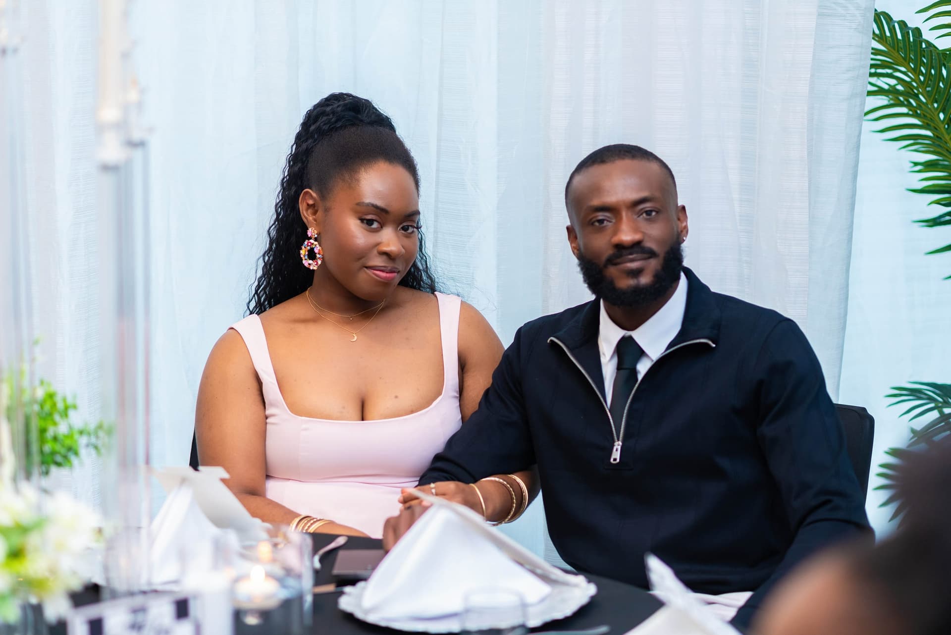 A pair at the far side of a table, she in blush and he in a dark zip-neck jacket, the room curtained in soft light behind.