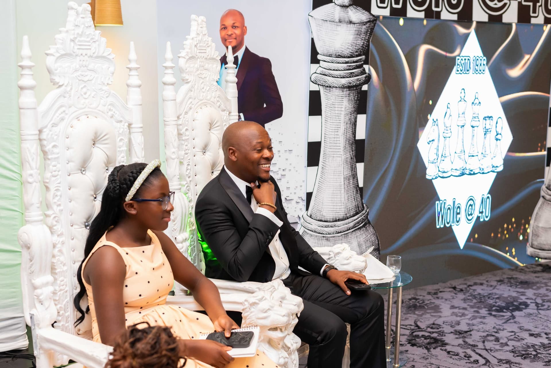 He takes one of two white ornate thrones set in front of the backdrop, his eldest daughter already seated beside him.